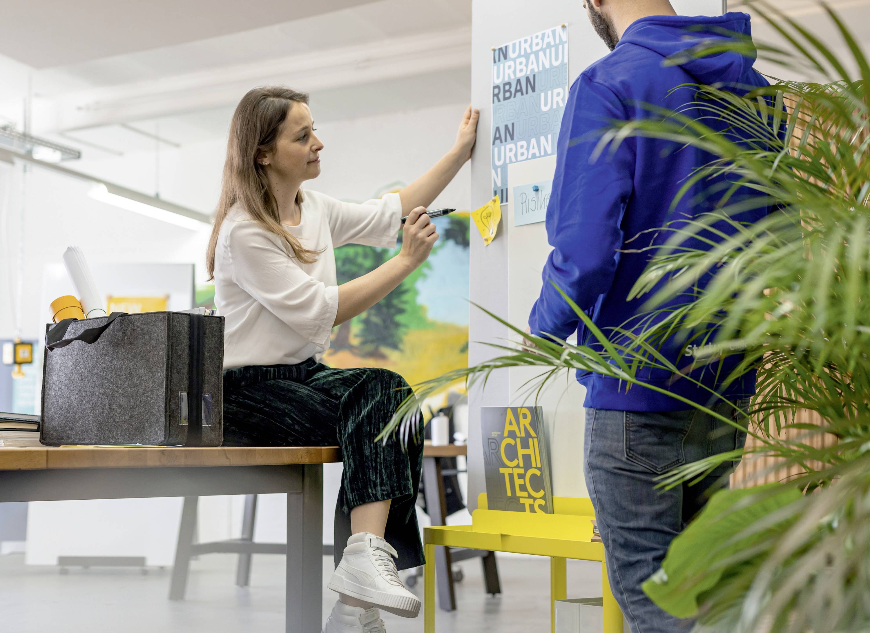 Žena sedí u stolu a spolu s mužem rozmisťuje plakáty na stěně. Diskutují o designu nebo architektuře.