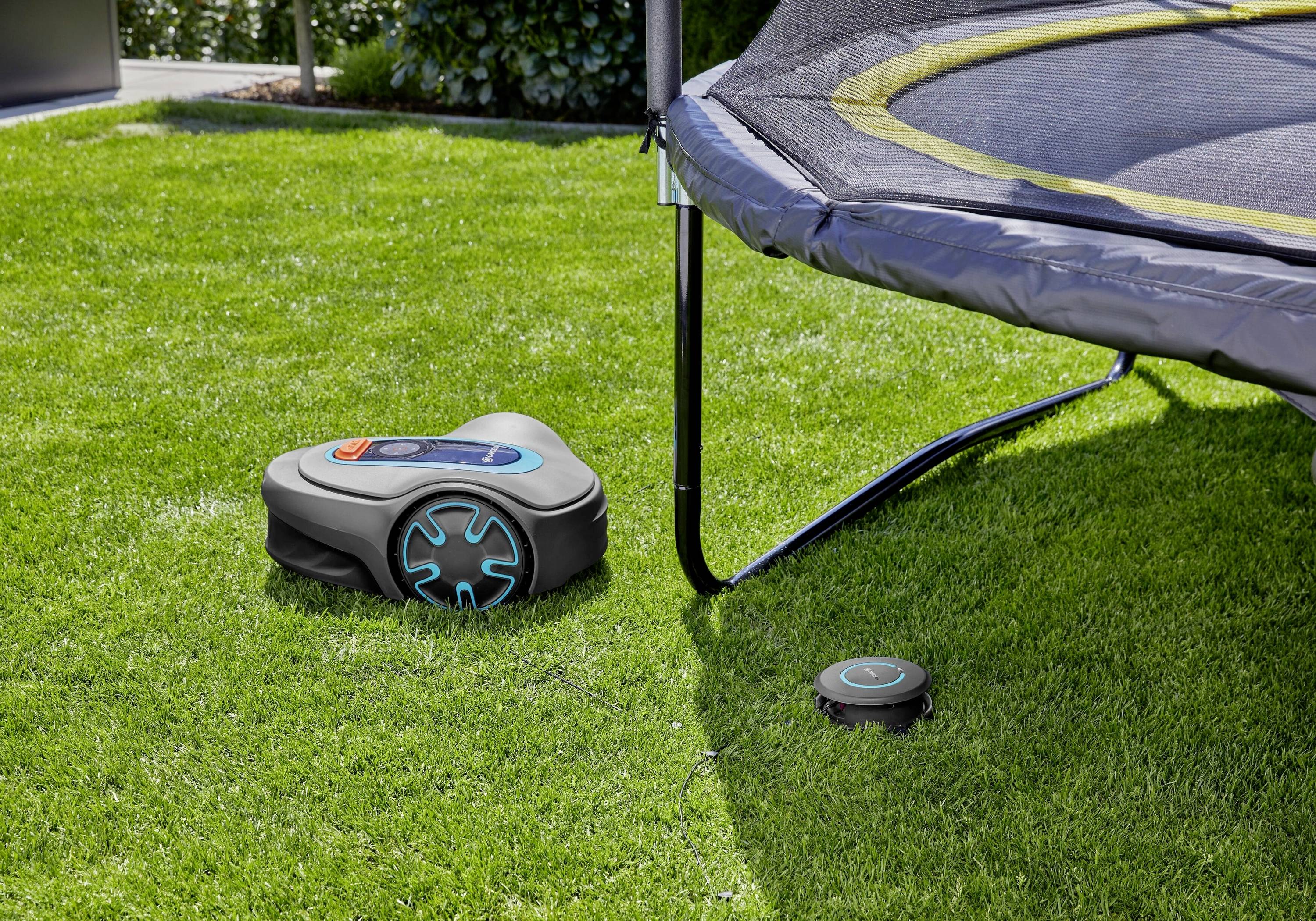Robotická sekačka seká trávu vedle trampolíny na zahradě. Malé zařízení leží na trávníku jako součást sekacího systému.