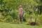 Muž přechází přes zelený trávník s navinutou zahradní hadicí v ruce, obklopen bujnou vegetací a stromy.