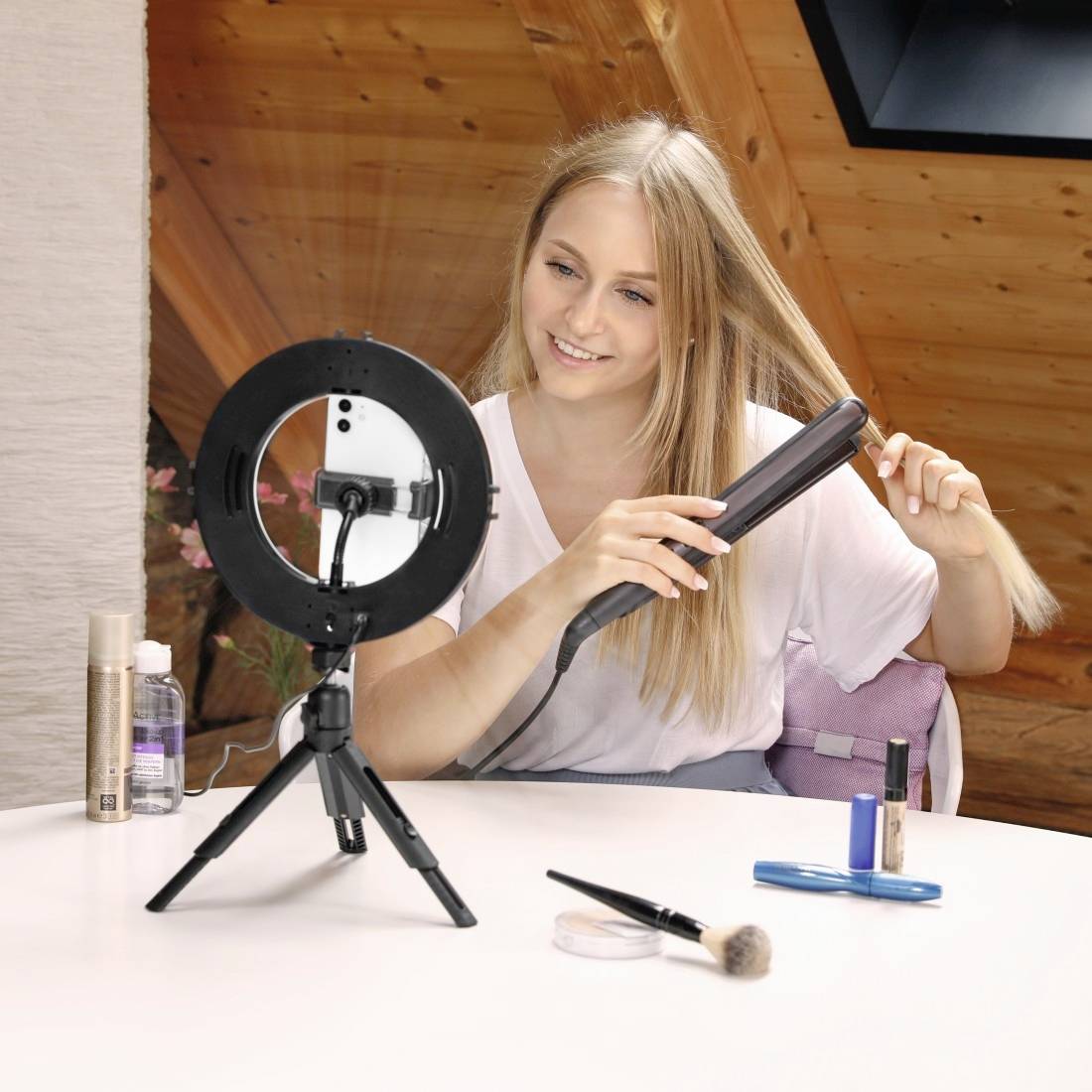Žena sedí u stolu a pomocí smartphonu umístěného na stativu s kruhovým osvětlením natáčí svůj make-up proces.