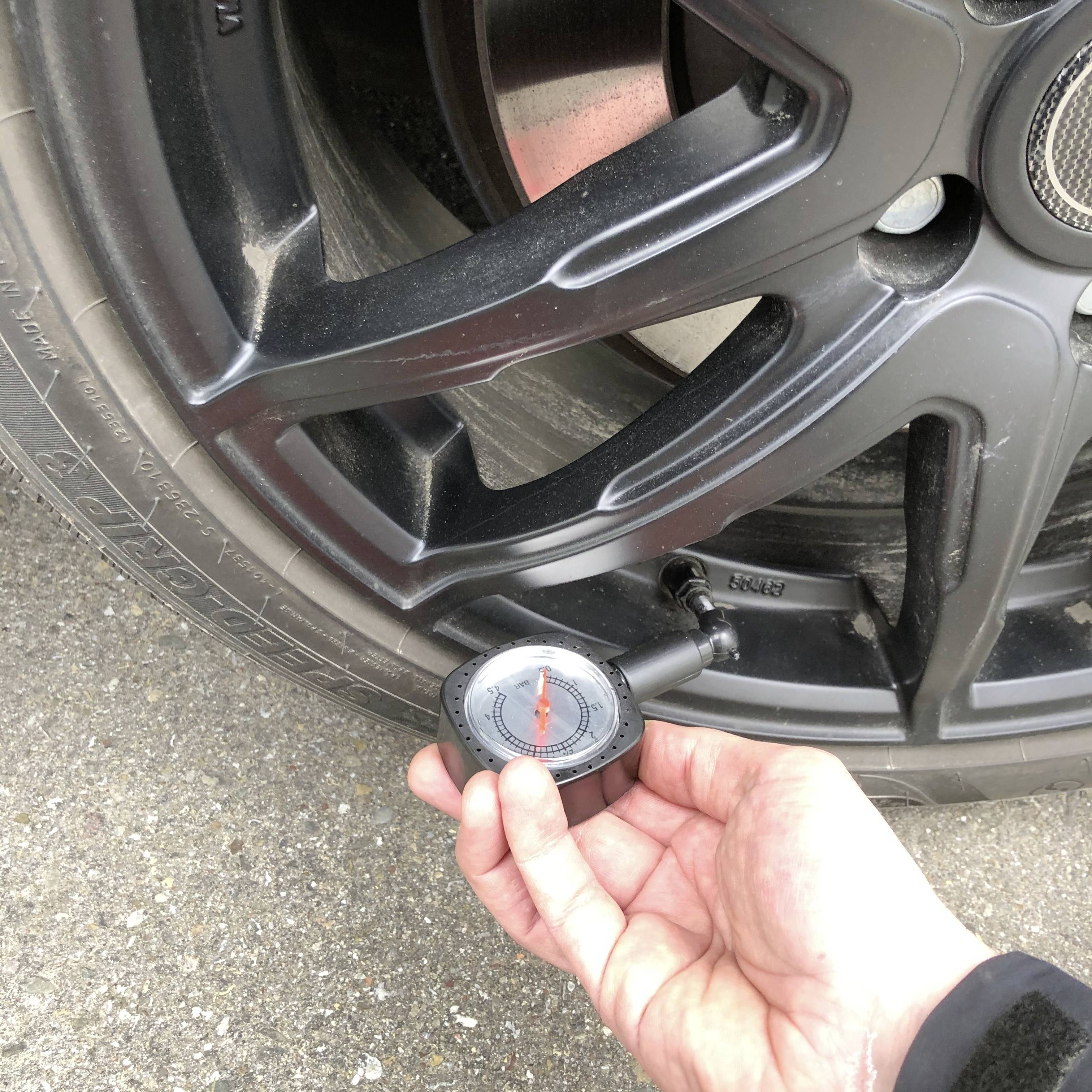 Ruka kontroluje tlak pneumatiky automobilu tlakoměrem. Pneumatika je černá a nachází se na asfaltu.