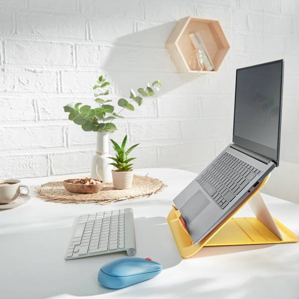 Laptop na žlutém stojanu stojí na psacím stole vedle rostliny, misky s ořechy a šálku.