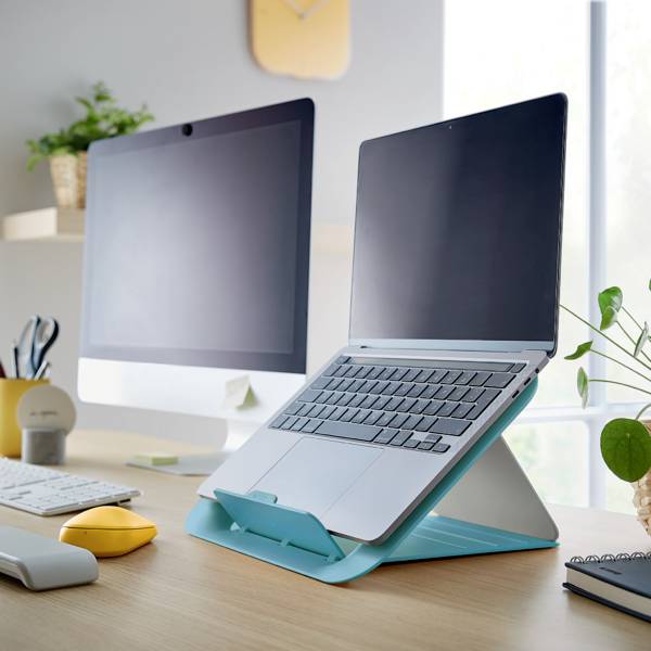 Laptop stojí šikmo na modrém stojanu vedle zapnutého stolního počítače v moderní kanceláři s rostlinami.