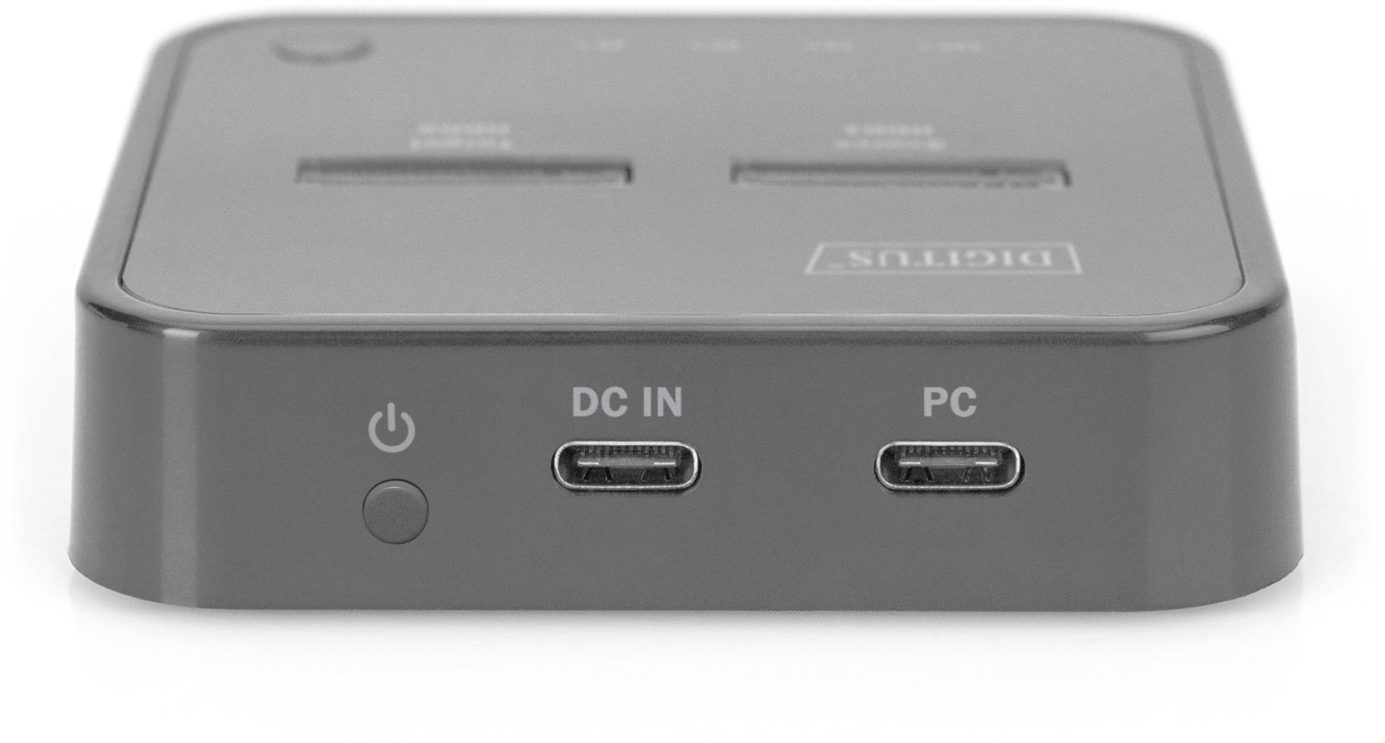 Šedý USB hub se dvěma USB-C porty, tlačítkem napájení a nápisy 'DC IN' a 'PC' na přední straně.