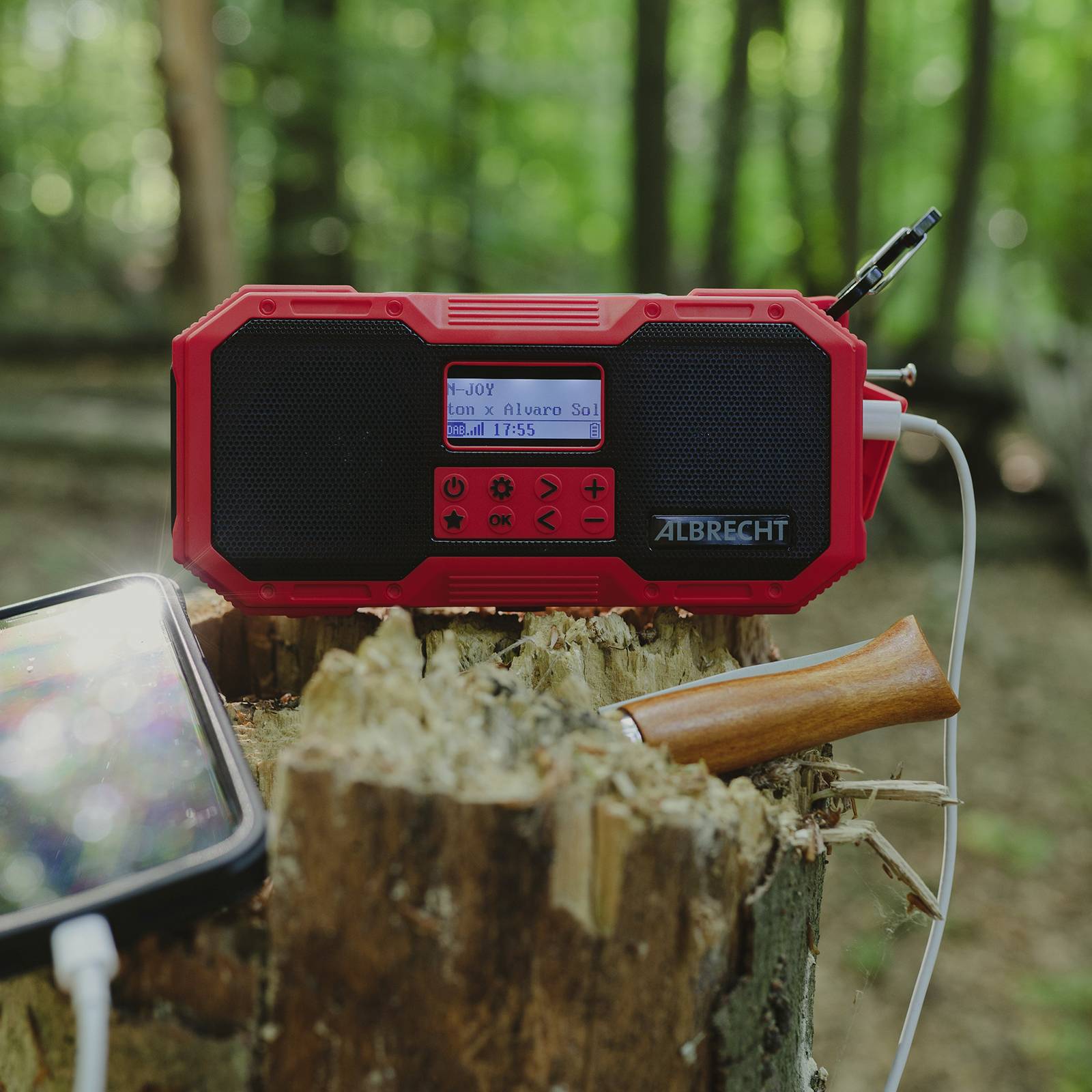 Červené přenosné rádio s displejem zobrazuje FM stanici '103.0'. Stojí na pařezu v lese. Vedle něj leží smartphone se sluchátky.