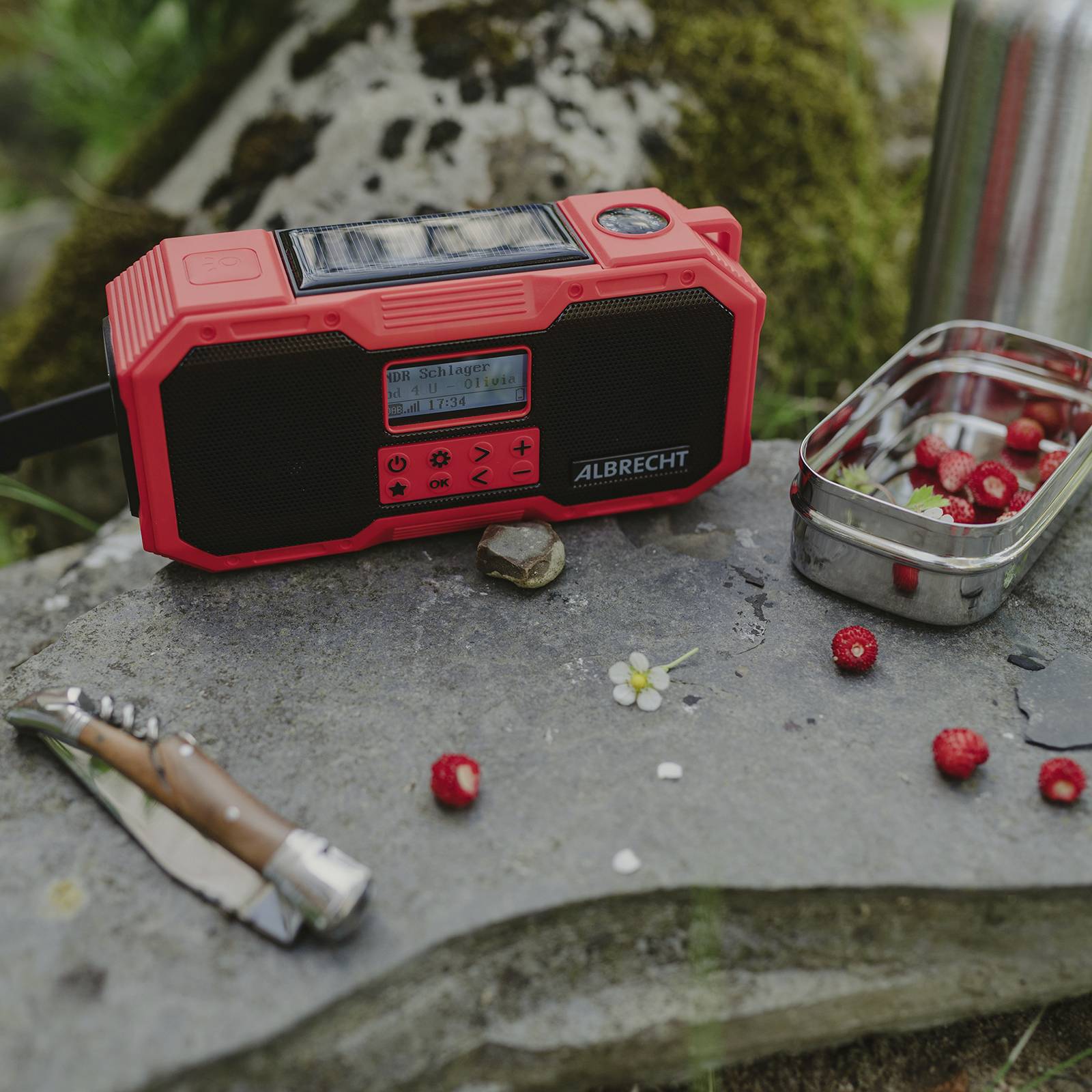 Červené přenosné rádio na kameni venku, vedle něj kapesní nůž a malá dóza s jahodami.