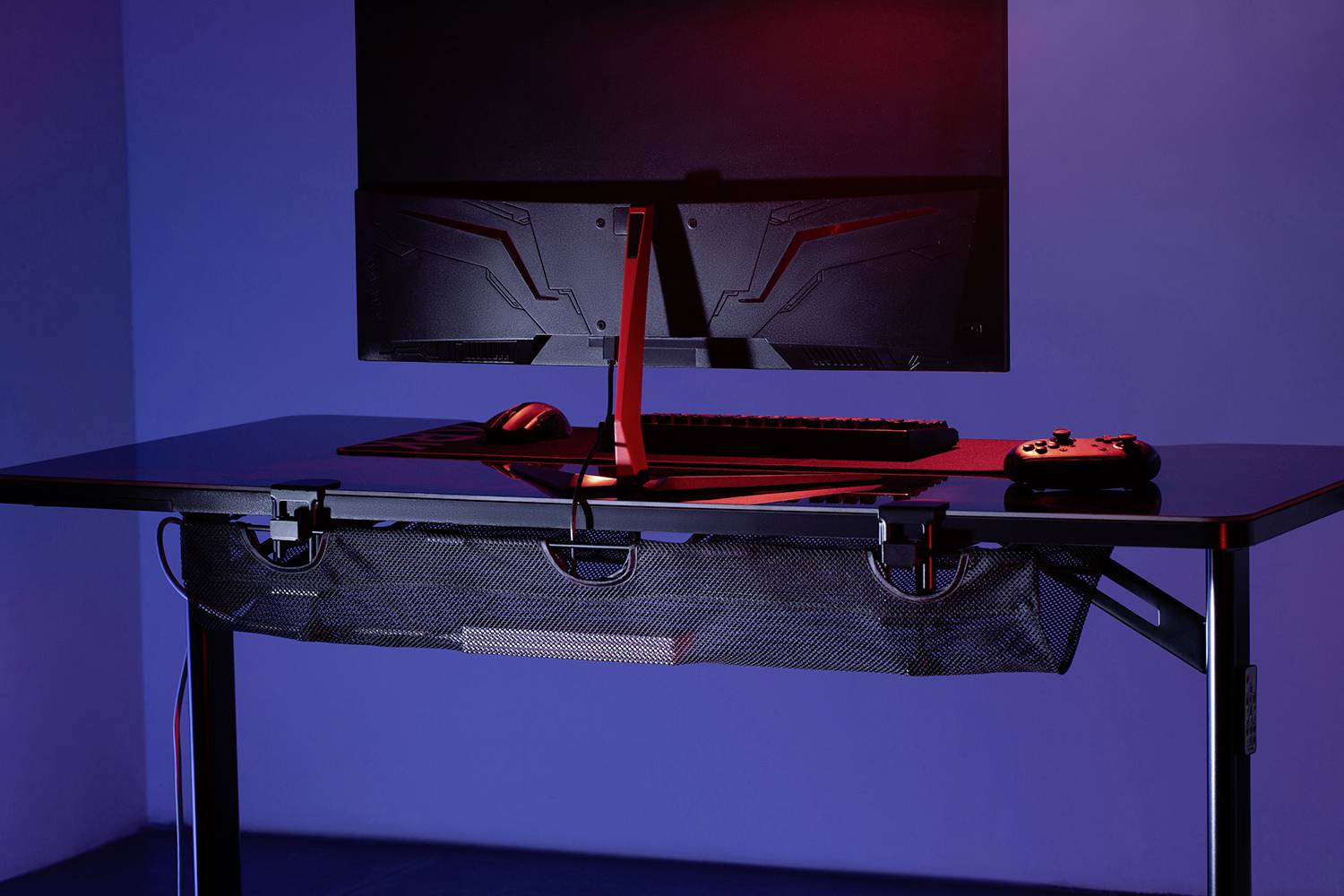 Moderní psací stůl s počítačovou obrazovkou, klávesnicí a herním ovladačem. Červené osvětlení dodává prostoru futuristickou atmosféru.