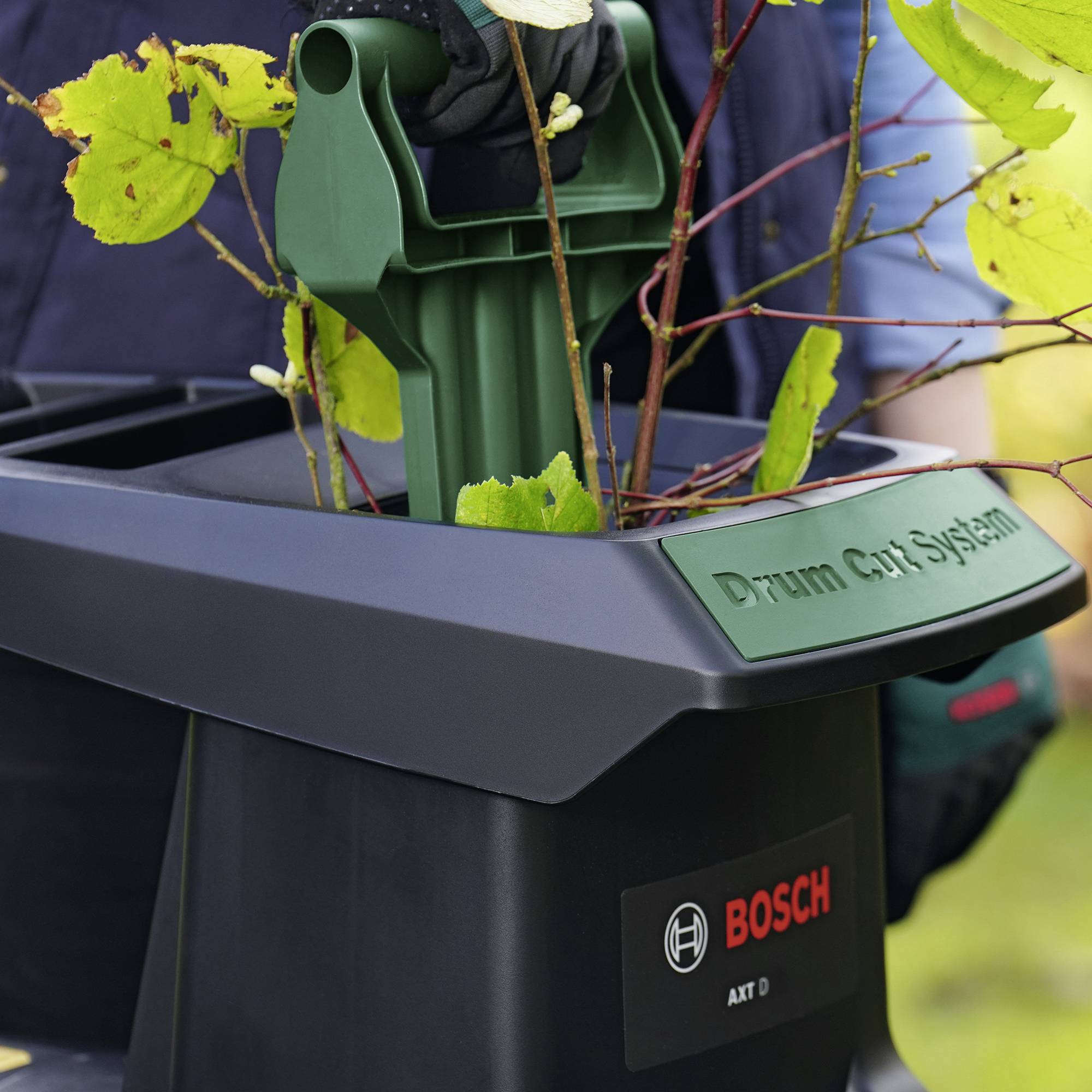 Detailní záběr černé stanice zahradního nářadí s nápisem 'Drum Cut System' a logem Bosch. Zelené zahradní zařízení s rostlinami na povrchu.