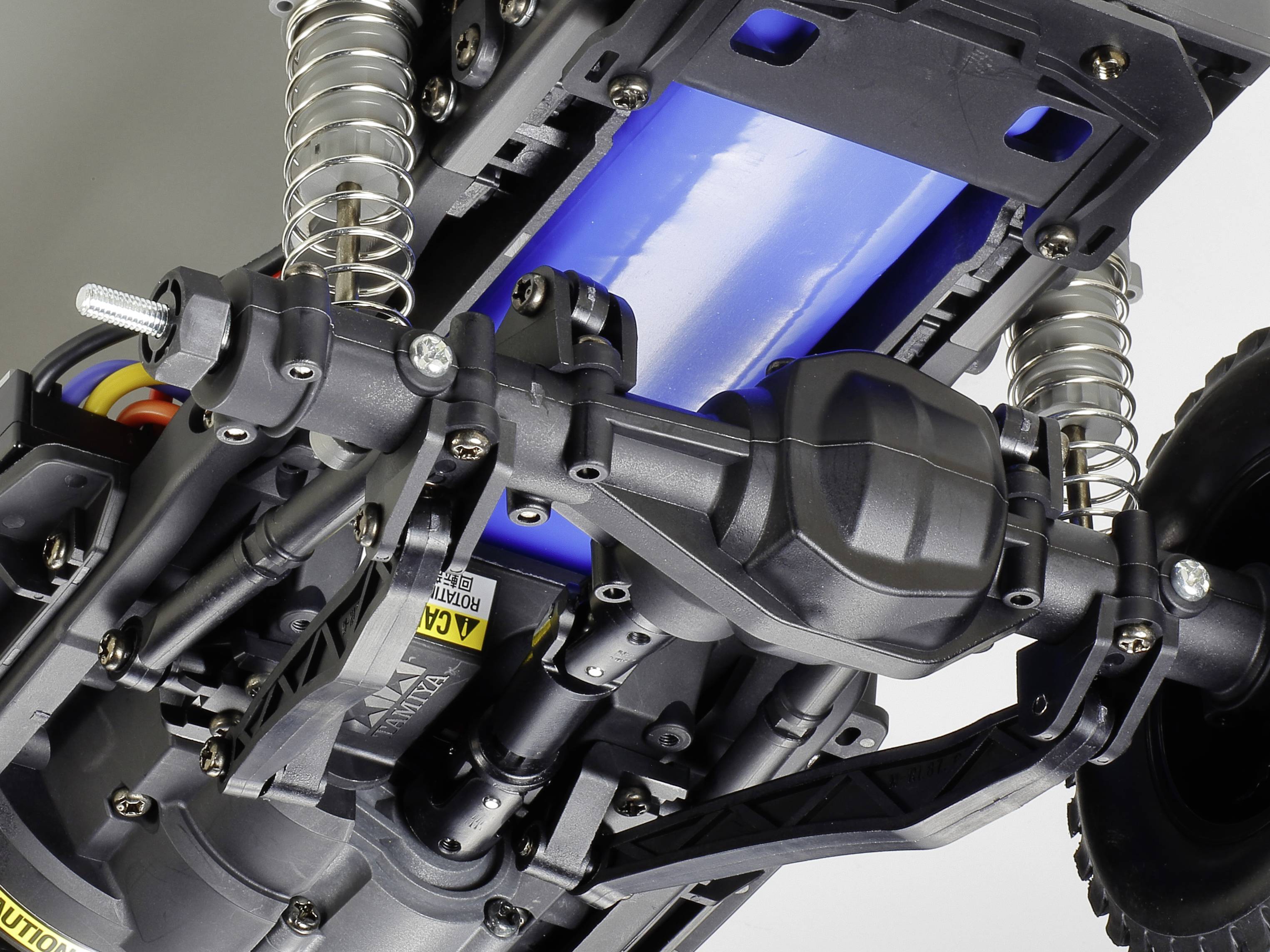 Detailní záběr modelu RC automobilu. Viditelné jsou zavěšení, převodovka a modrá baterie. Zaměření je na techniku a mechanické detaily.