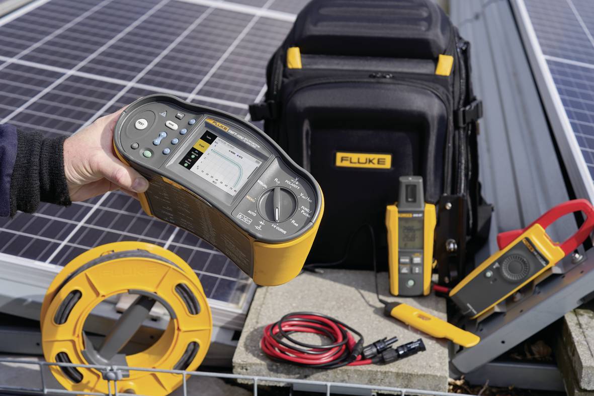 Přístroj pro měření elektrických parametrů je držen osobou, v pozadí jsou další nástroje a solární panely.