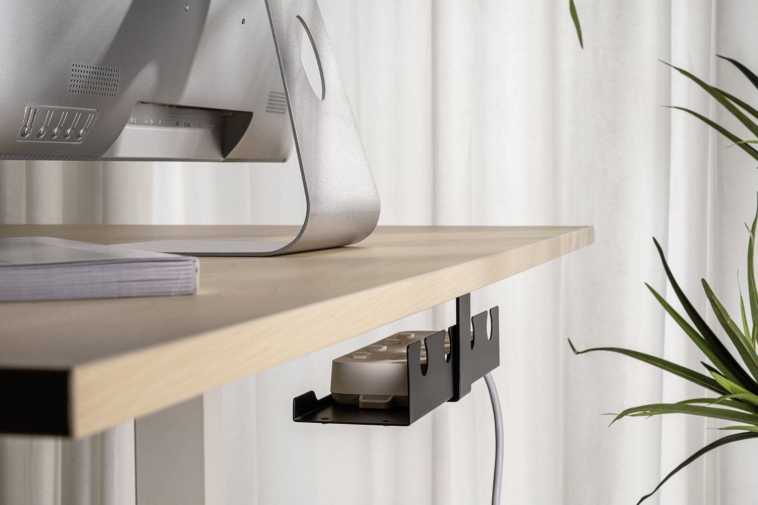 Počítačový monitor stojí na dřevěném psacím stole. Pod ním je upevněn černý organizér kabelů s několika kabely. V pozadí jsou umístěny rostliny.