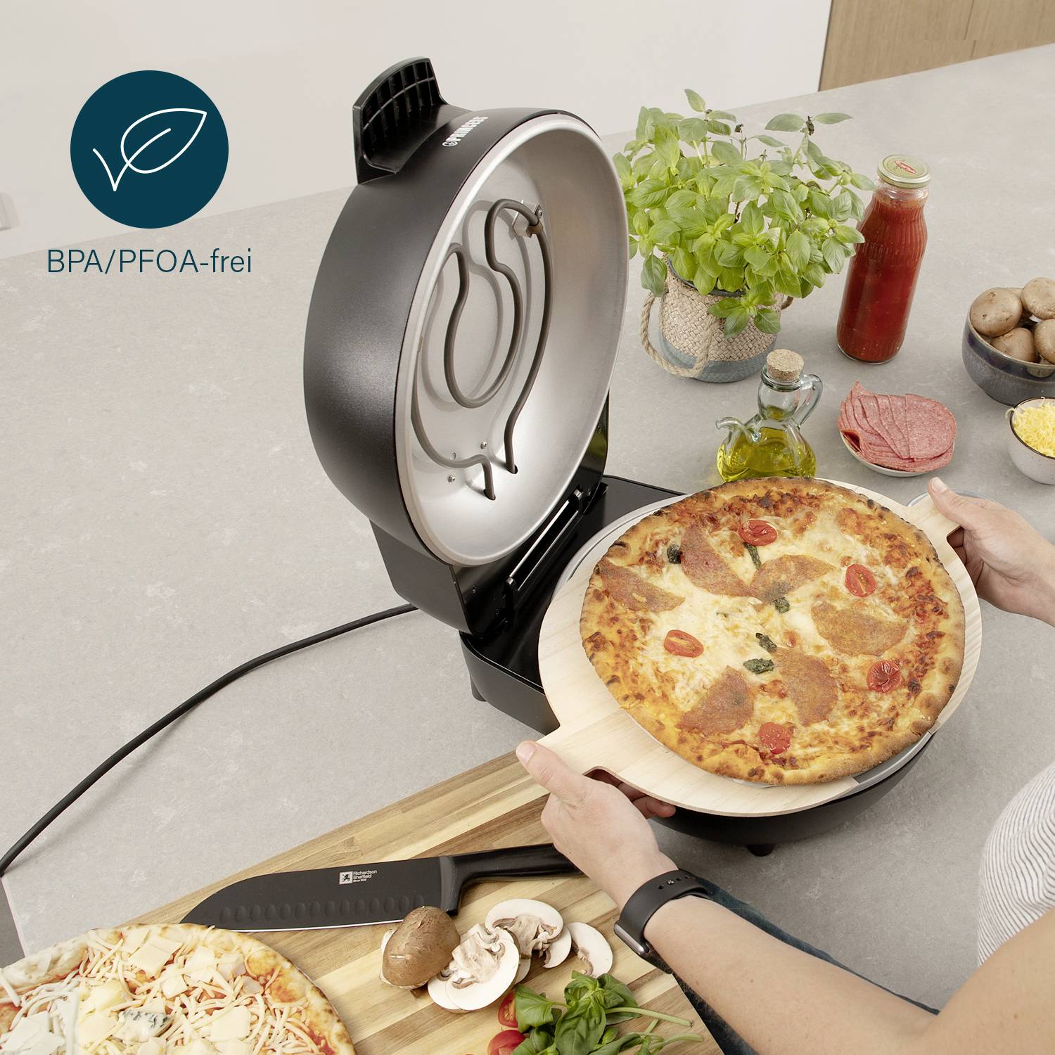 Osoba peče pizzu v elektrickém pizzovači. Na stole jsou ingredience jako rajčata, omáčka a houby. Štítek ukazuje 'bez BPA/PFOA'.