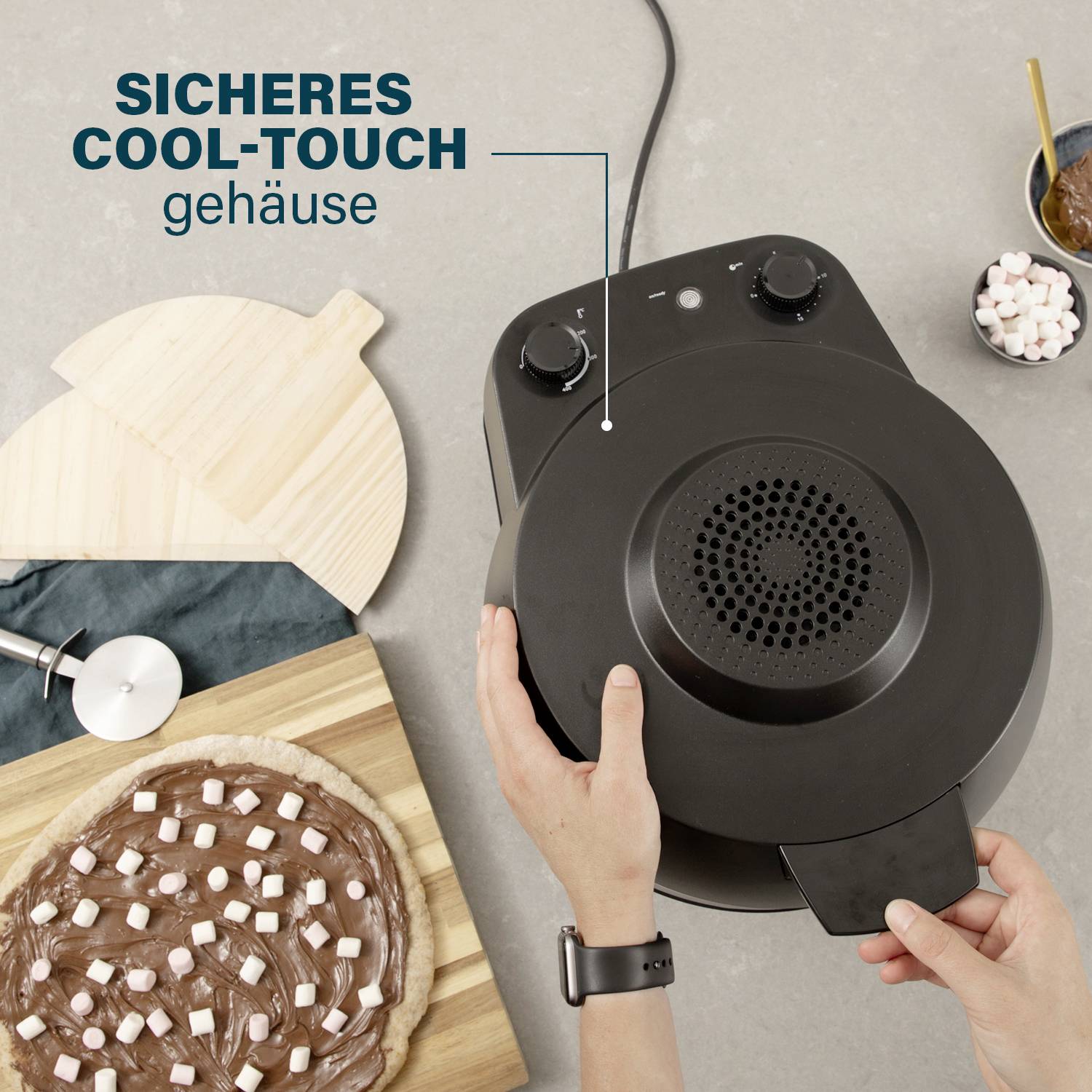 Dvě ruce obsluhují černý kuchyňský robot s nápisem "Bezpečné Cool-Touch pouzdro". Vedle leží pizza s marshmallow.