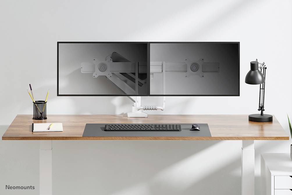 Dřevěný stůl s klávesnicí, myší, dvěma vedle sebe umístěnými monitory a černou stolní lampou. Vlevo stojí šálek.