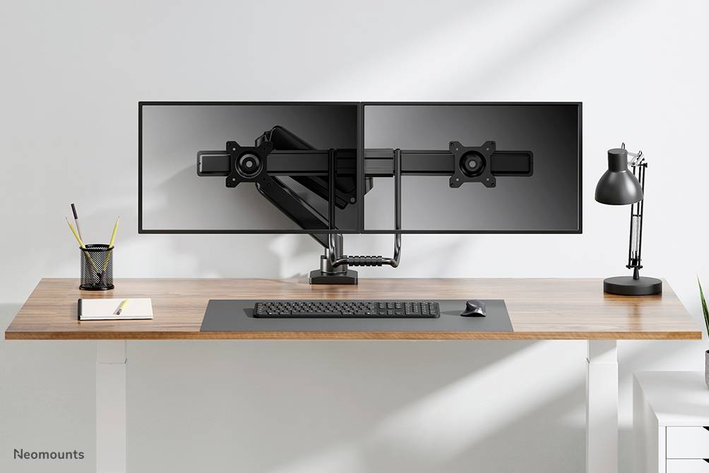 Dva prázdné držáky monitorů na psacím stole s lampou, klávesnicí a notebookem. Jednoduché, moderní kancelářské uspořádání.
