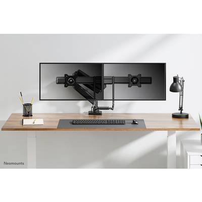 Dva prázdné držáky monitorů na psacím stole s lampou, klávesnicí a notebookem. Jednoduché, moderní kancelářské uspořádání.