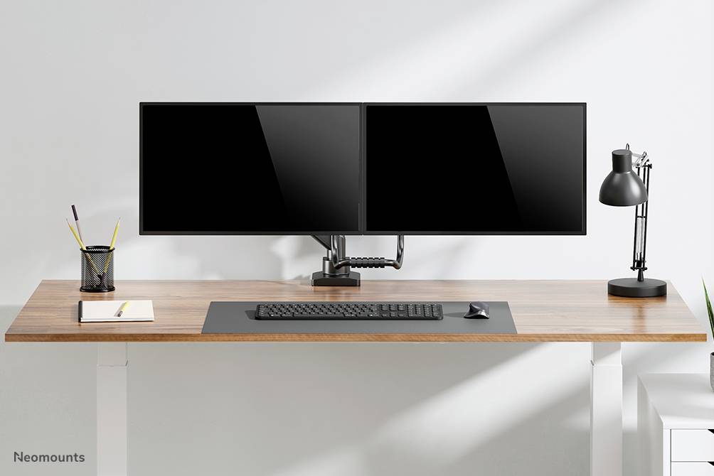 Uklizený pracovní stůl se dvěma monitory, klávesnicí, psacími potřebami a lampou. Minimalistický, moderní design.