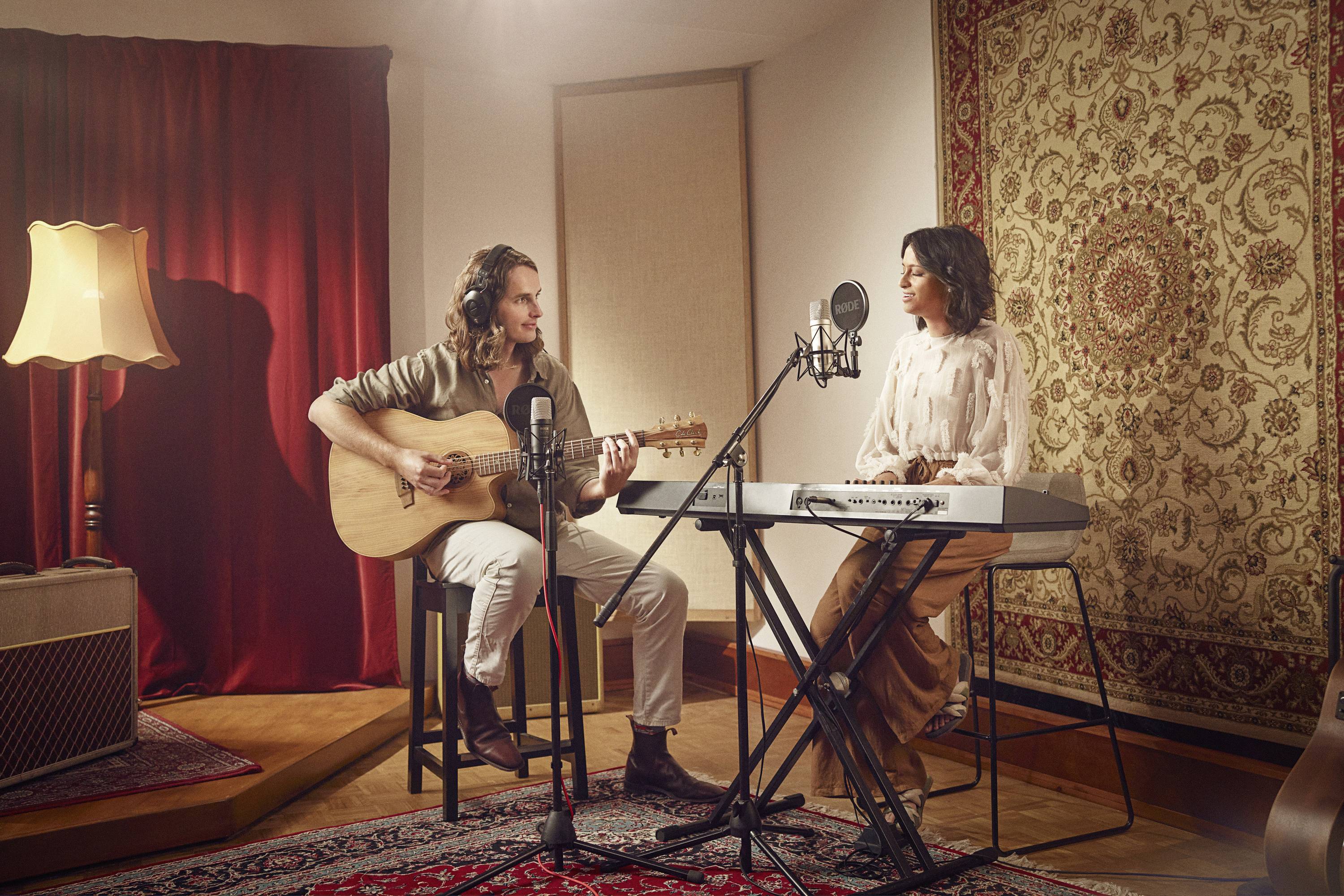 Dvě osoby hrají hudbu ve studiu. Vlevo někdo hraje na kytaru, vpravo někdo hraje na klávesy. Mikrofony jsou postavené.