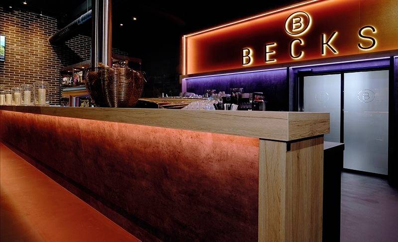 Bar s osvětleným pultem a cedulí s nápisem 'BECKS'. Atmosféra je moderní a stylová.
