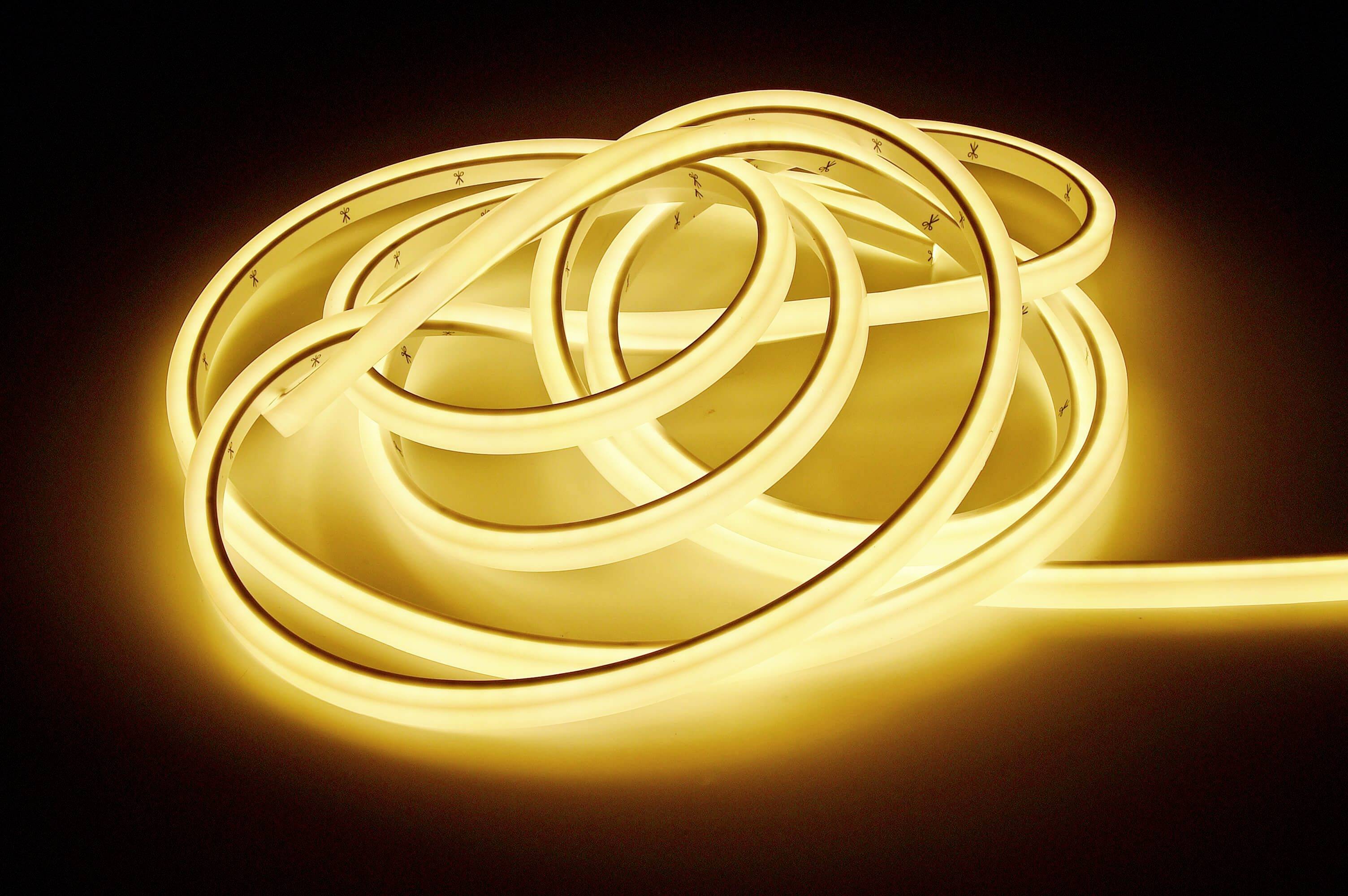 Úzký, jasně žlutý LED pásek ležící v nepravidelné smyčce, který vyzařuje teplé světlo v tmavé místnosti.