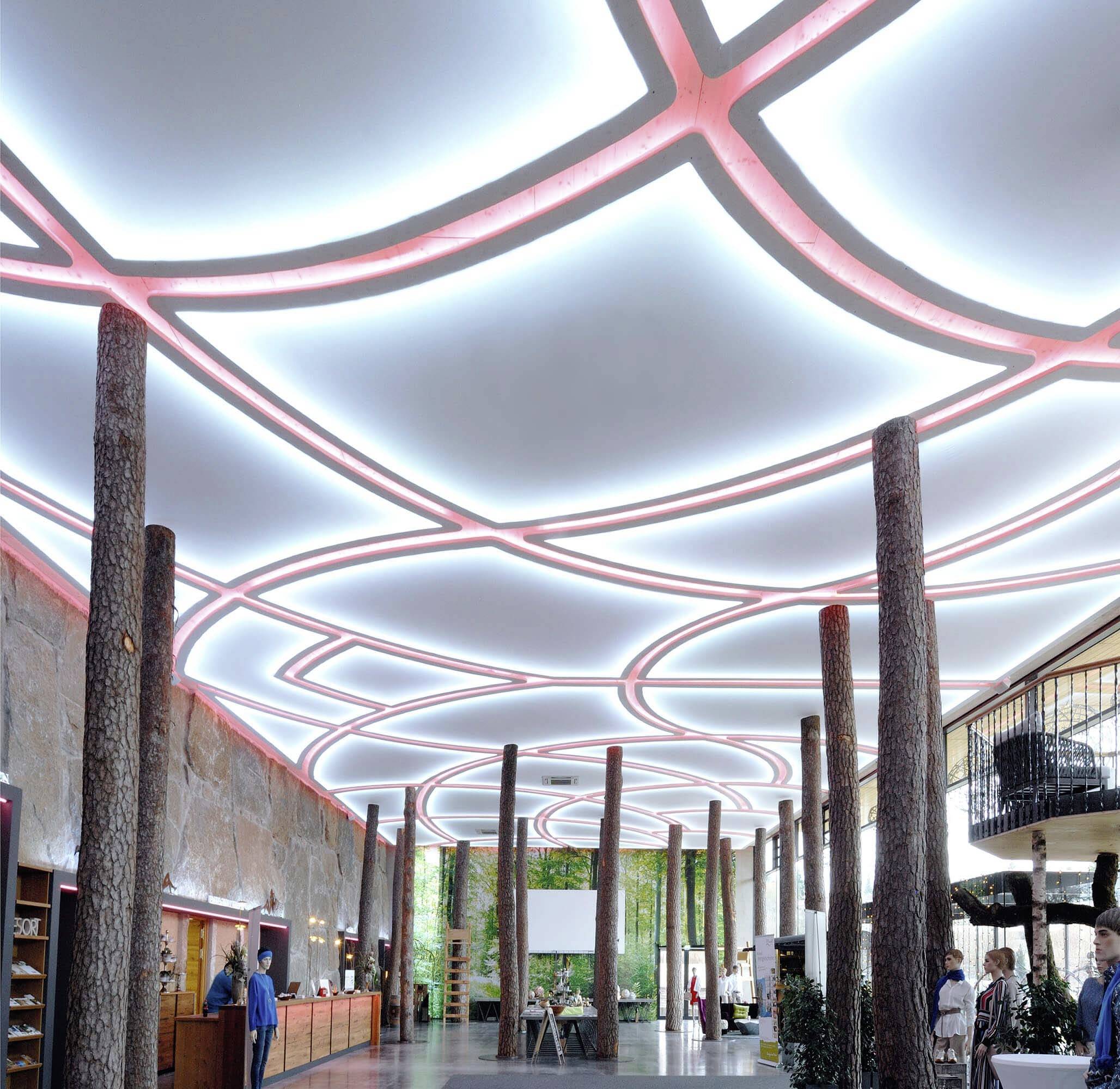 Hotelová lobby s moderním geometrickým osvětleným stropním vzorem z osvícených linií, zdobená sloupy ve tvaru kmenů stromů.