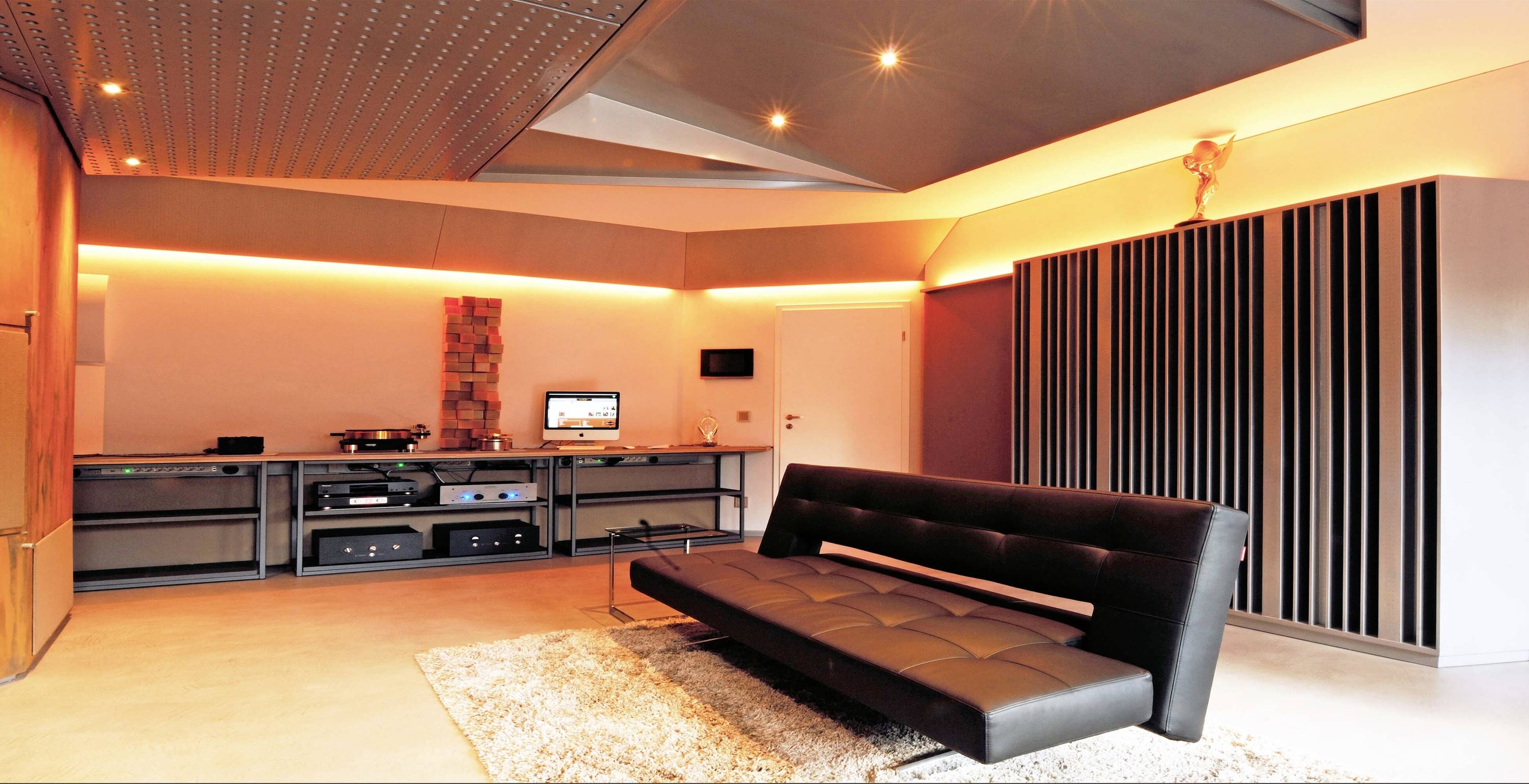 Moderní obývací pokoj s černou pohovkou, světlým kobercem a zábavním systémem. Stěny jsou vybaveny nepřímým osvětlením.