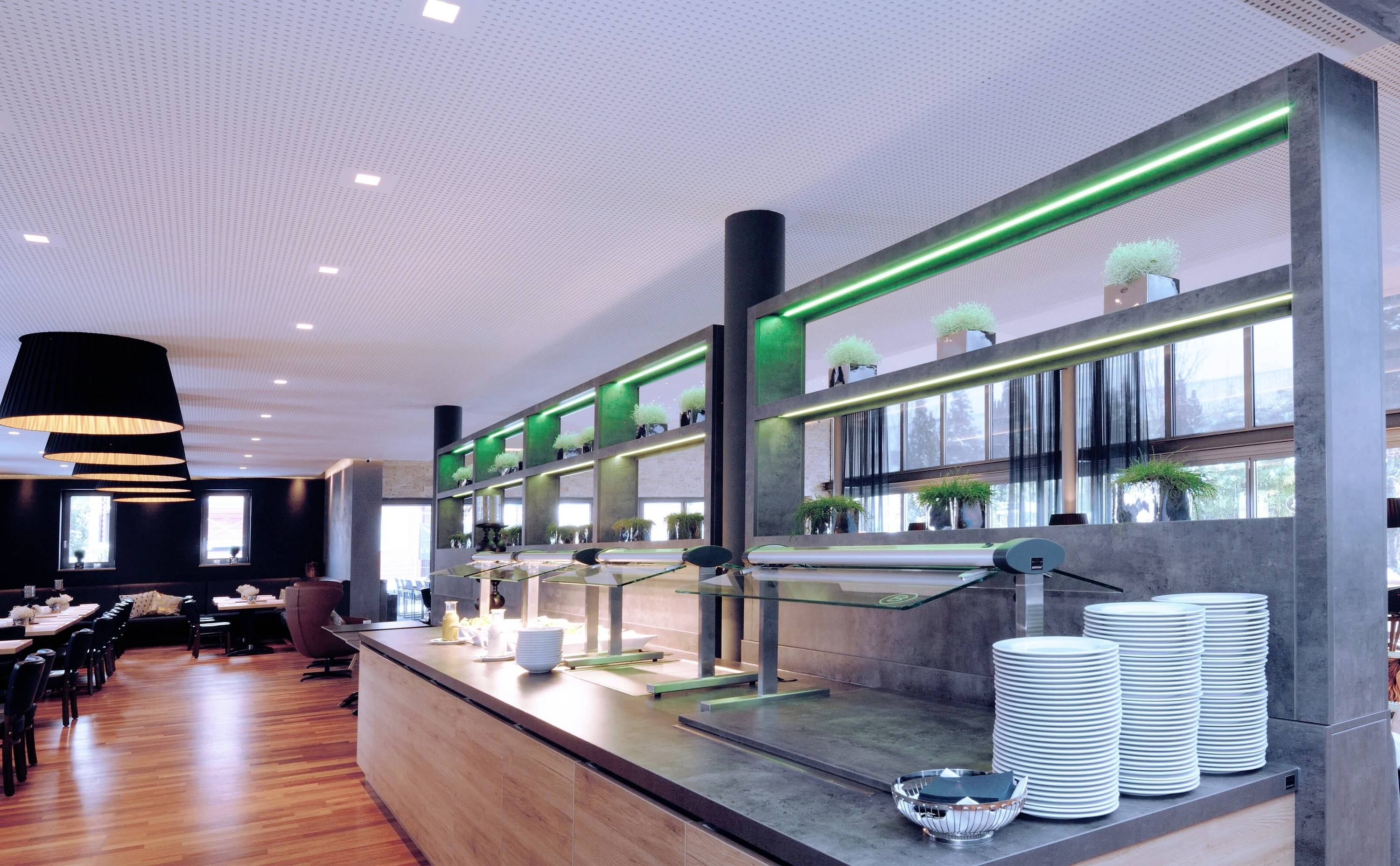 Moderní restaurace s dlouhým pultem, zelenými světly a naskládaným nádobím. V pozadí jsou vidět prostřené stoly.