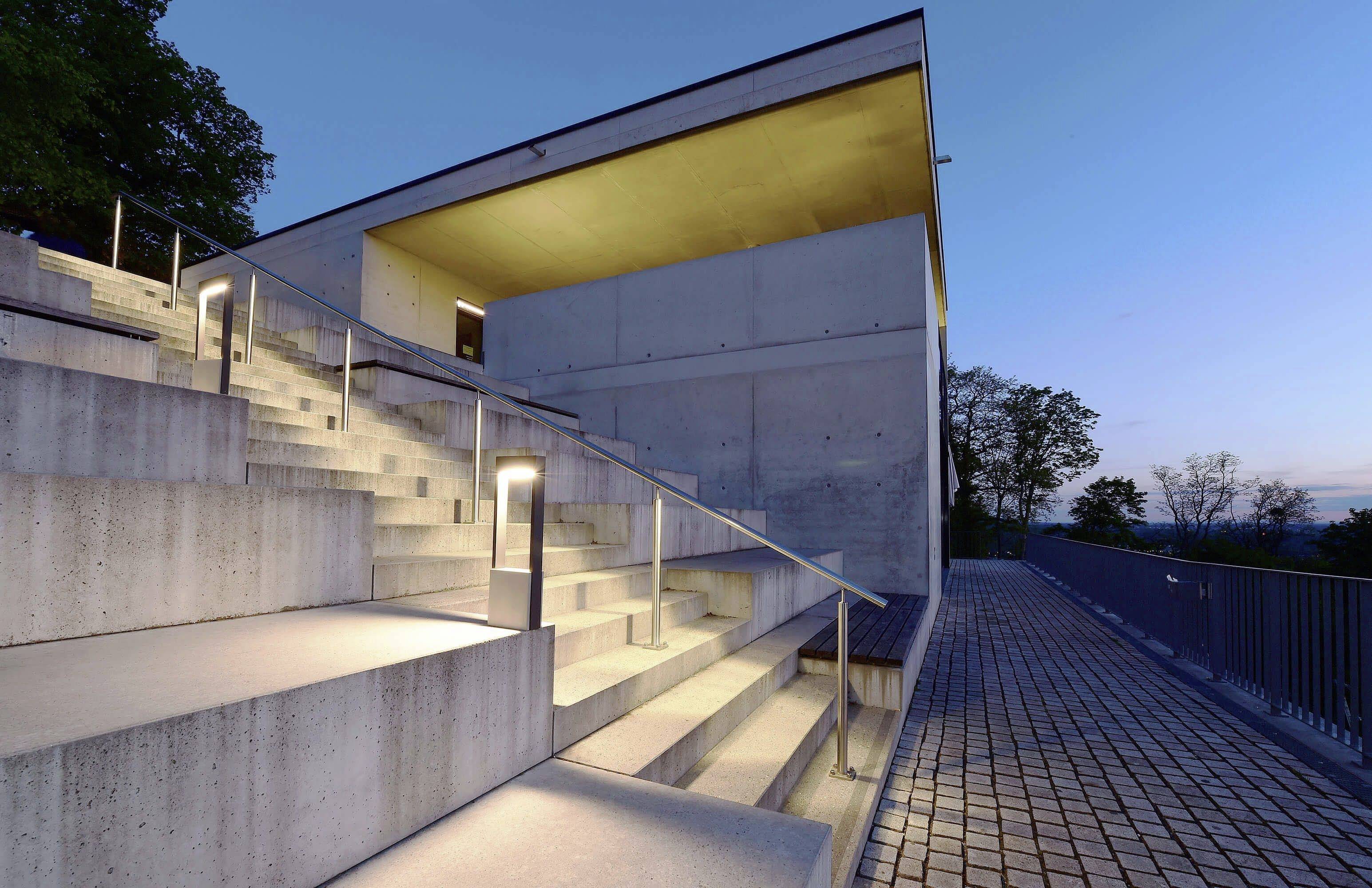 Moderní budova s osvětlenými schody a zábradlím za soumraku. Dominuje geometrický styl a čistá architektura.