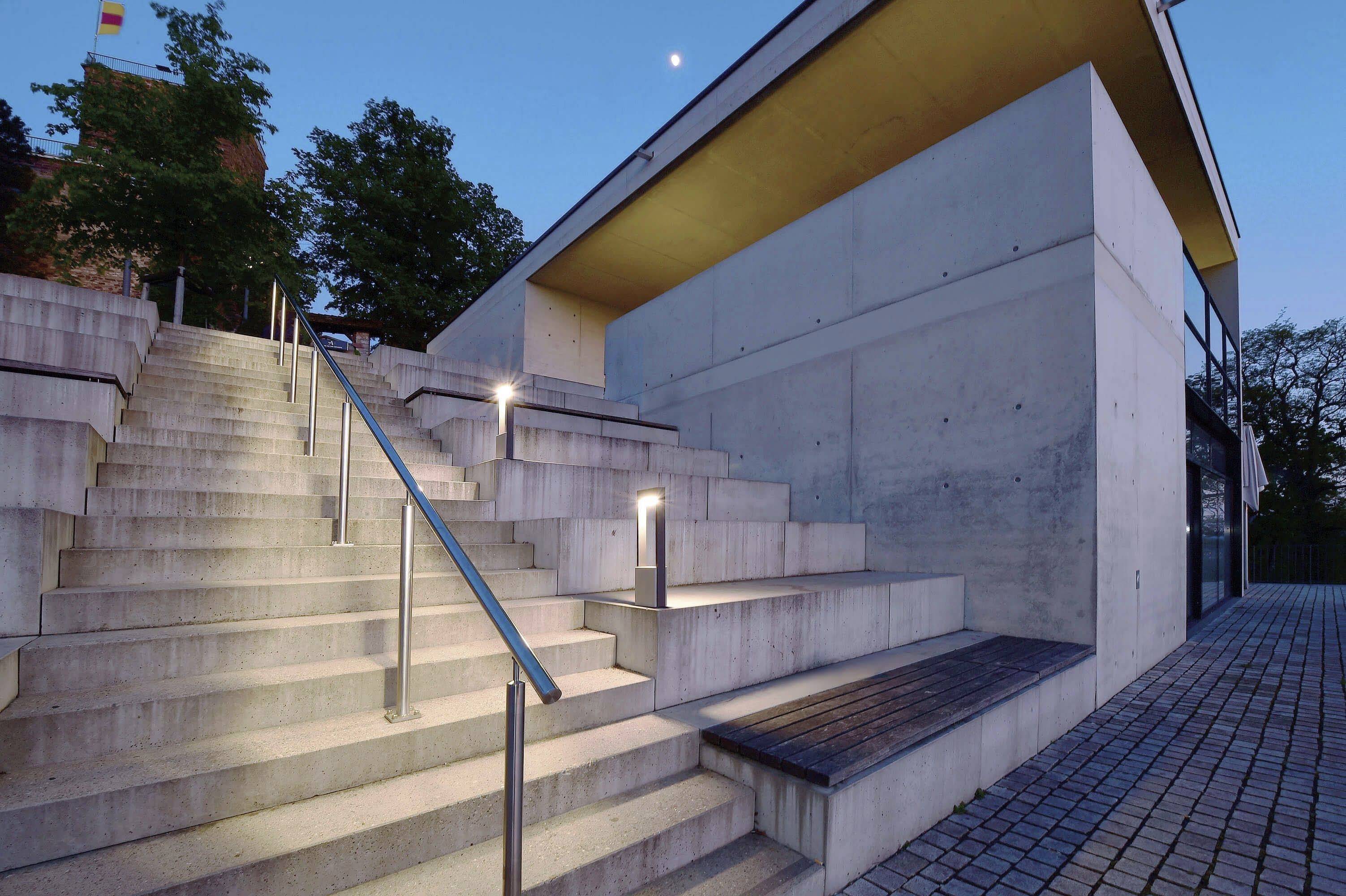 Moderní vstup do budovy s osvětlenými betonovými schody a zábradlím, pod nimi dlážděná cesta; obklopeno stromy za soumraku.