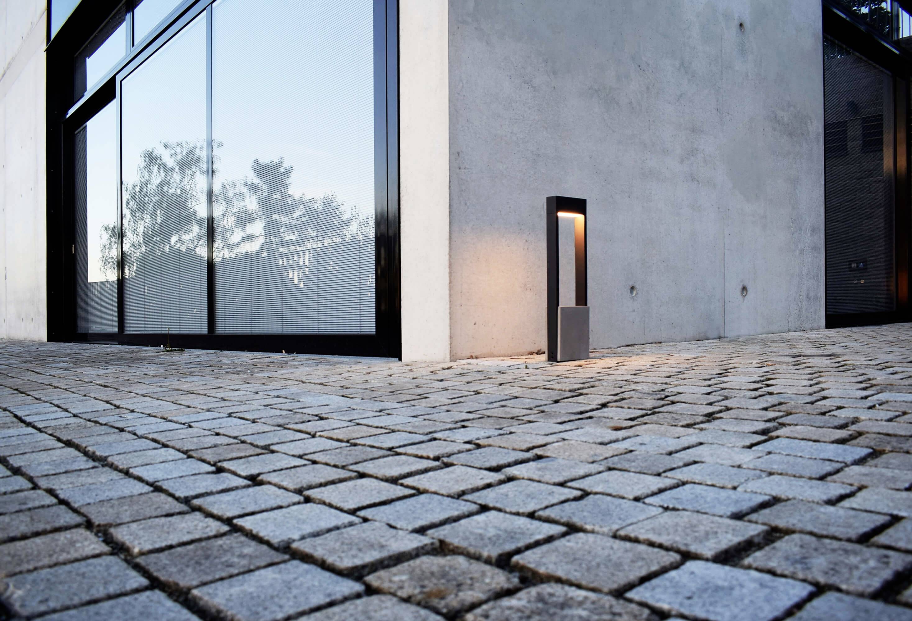 Moderní budova s velkými prosklenými okny, obklopená dlážděnou plochou. Venkovní osvětlení stojí vedle zdi.