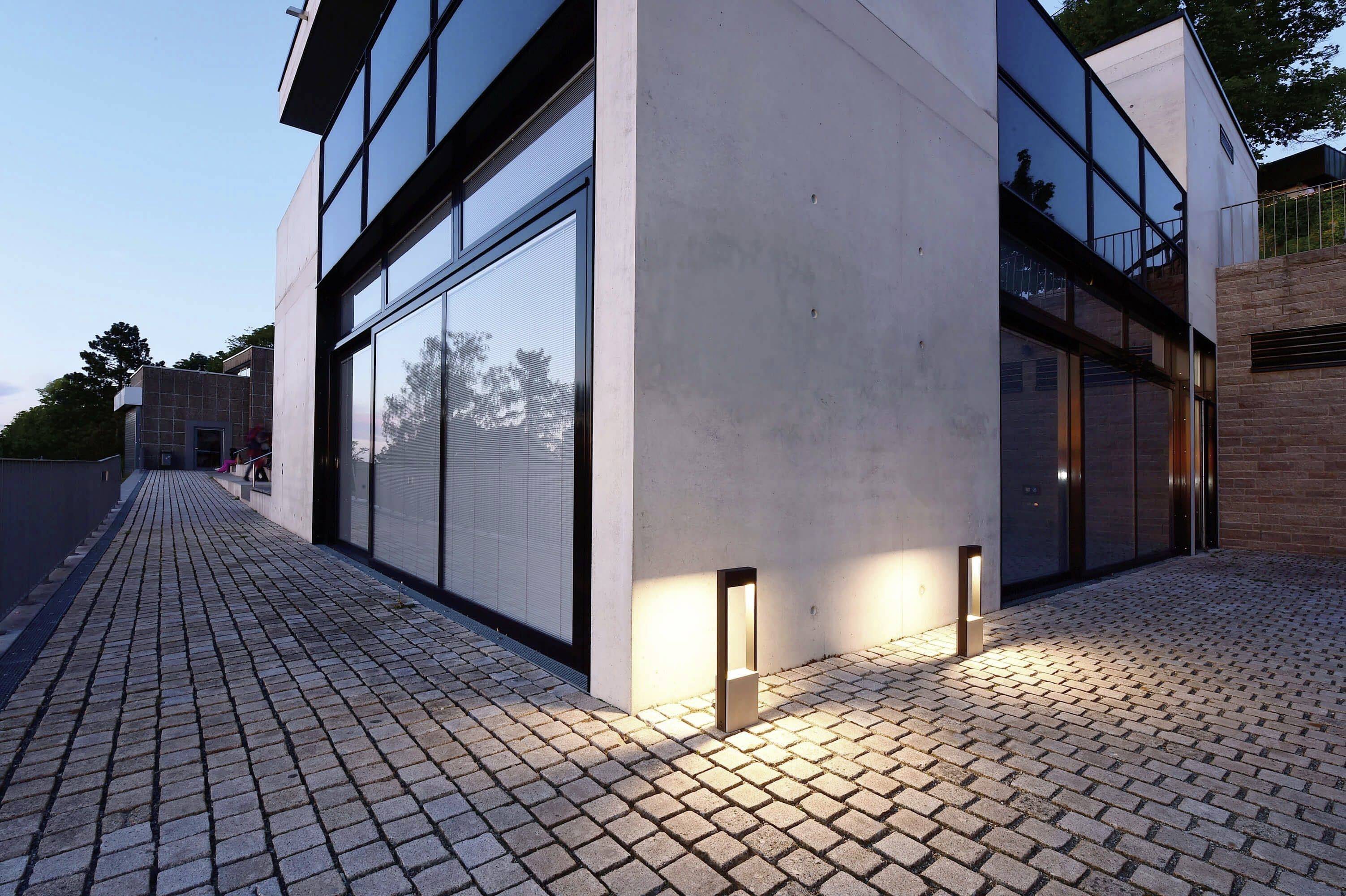 Moderní budova s velkými okny, obklopená dlažebními kostkami. Večerní scéna s osvětlením exteriéru.