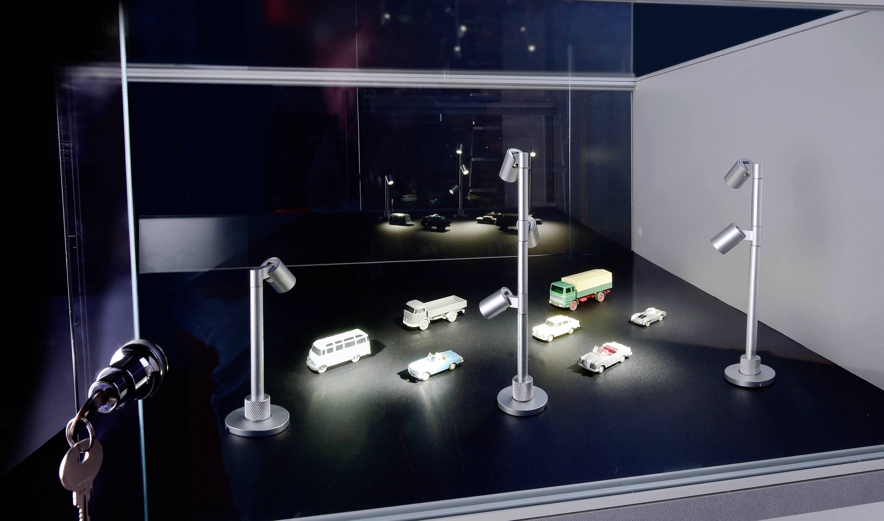 Miniaturní scéna s několika osvětlenými modelovými vozidly na černé ploše, obklopená malými pouličními lampami ve výstavním prostoru.