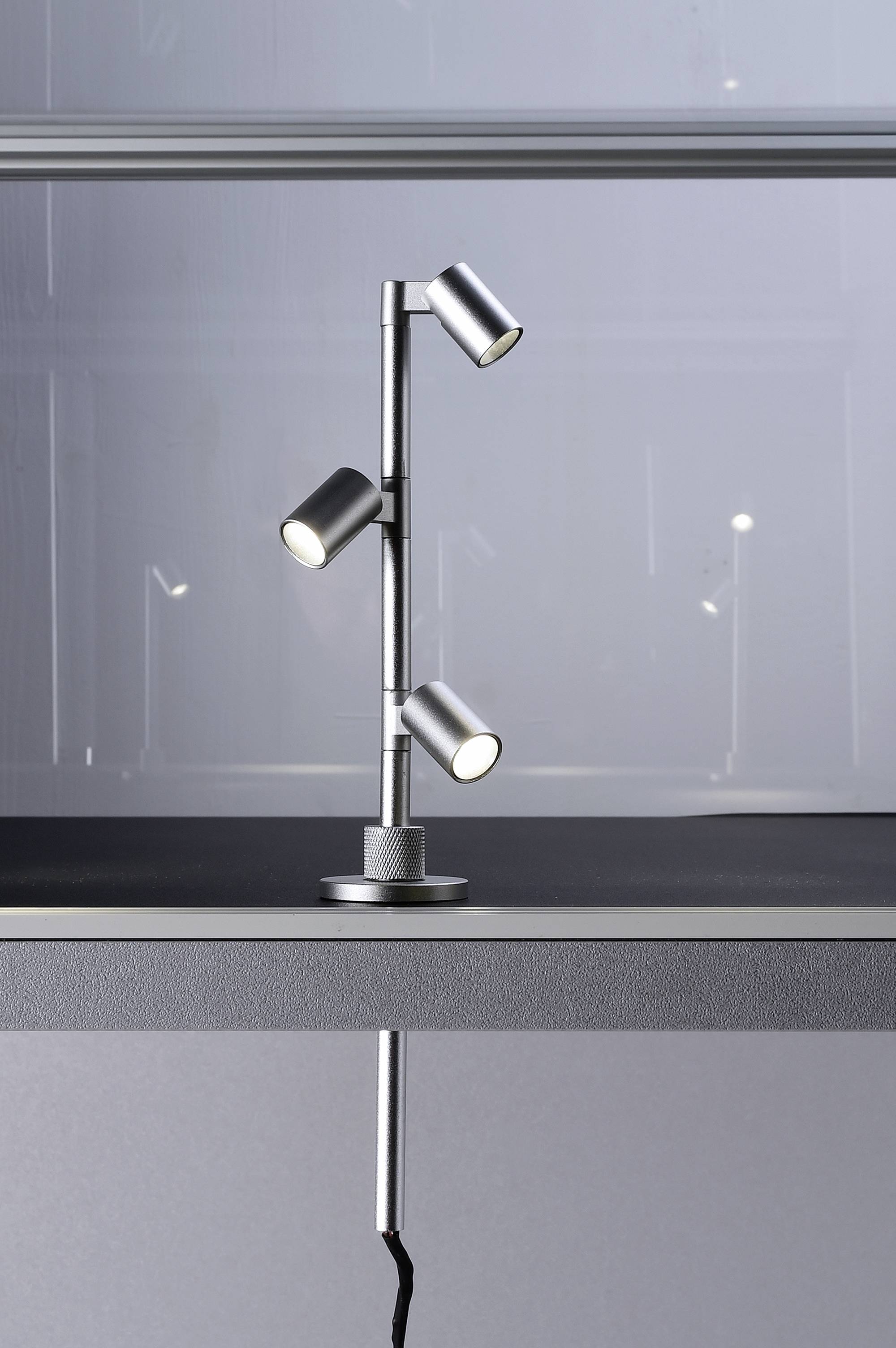 Moderní stolní lampa se třemi nastavitelnými LED světly na kovovém podstavci v minimalistickém prostoru.