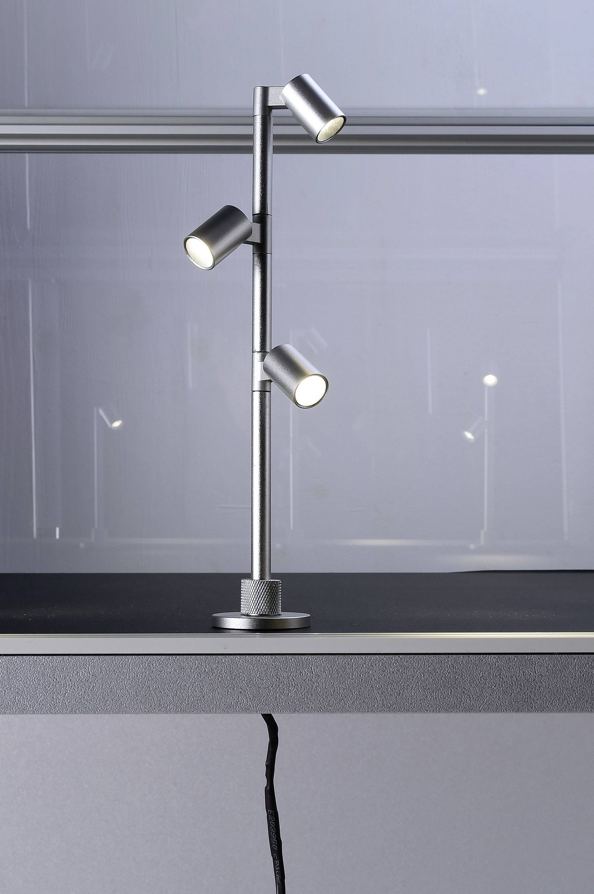 Tříramenná stříbrná stolní lampa na stole, která rovnoměrně osvětluje pracovní plochu. Čistý, moderní design.