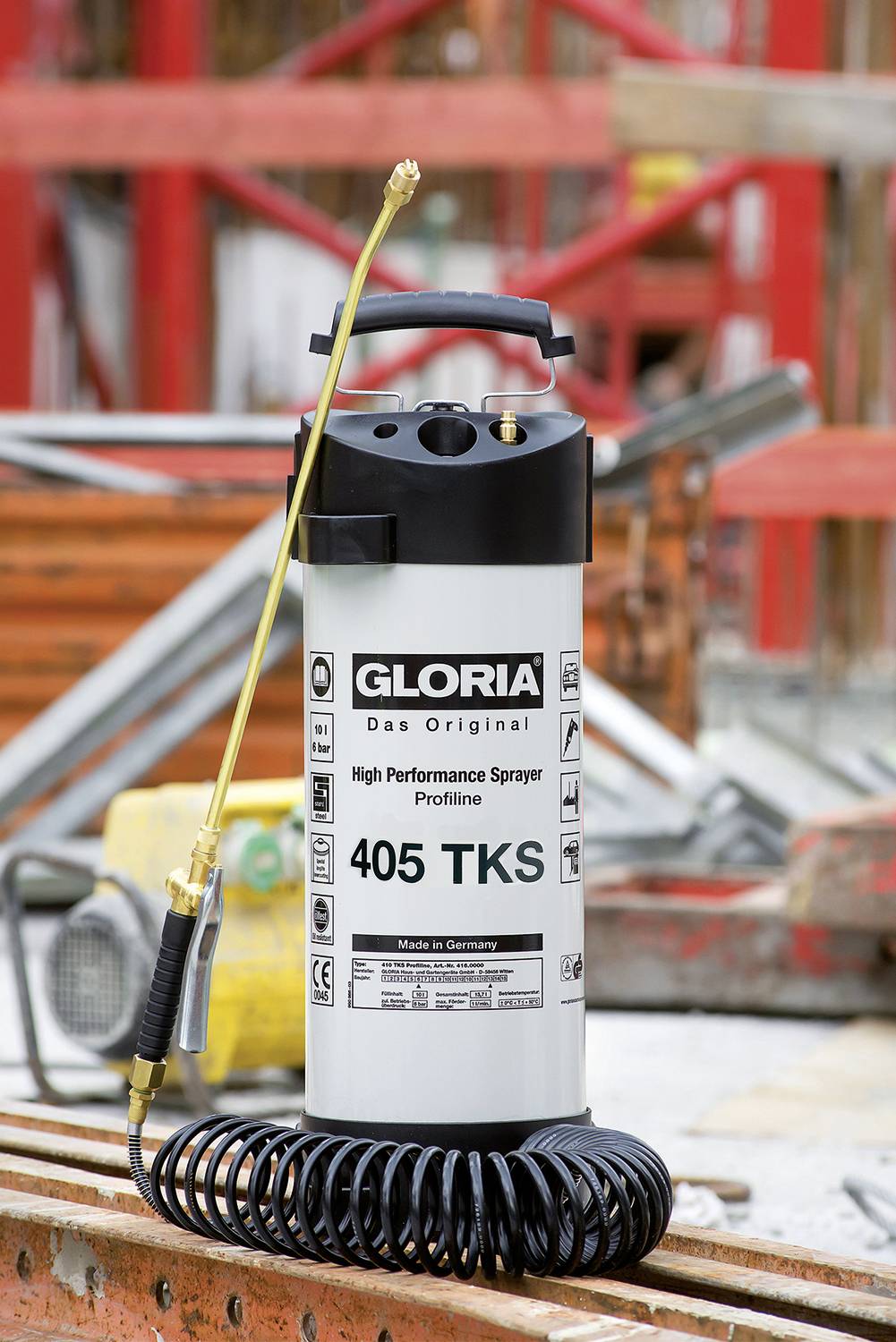 Vysokovýkonný postřikovač s nápisem 'Gloria 405 TKS' stojí před staveništěm. Má zlatý postřikovací nástavec a spirálovou smyčku.