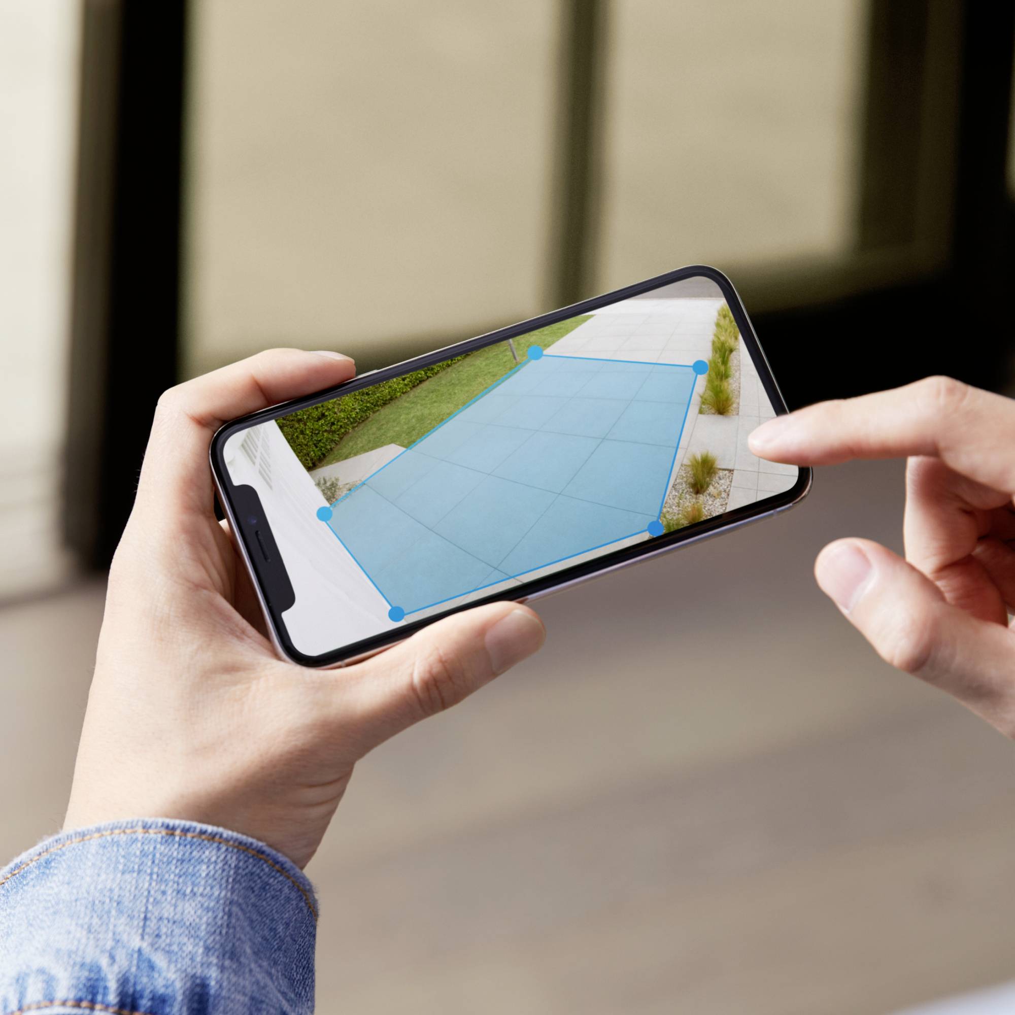 Osoba drží smartphone, který zobrazuje AR aplikaci. Aplikace měří obdélník na podlaze, pravděpodobně pro domácí kutilství.