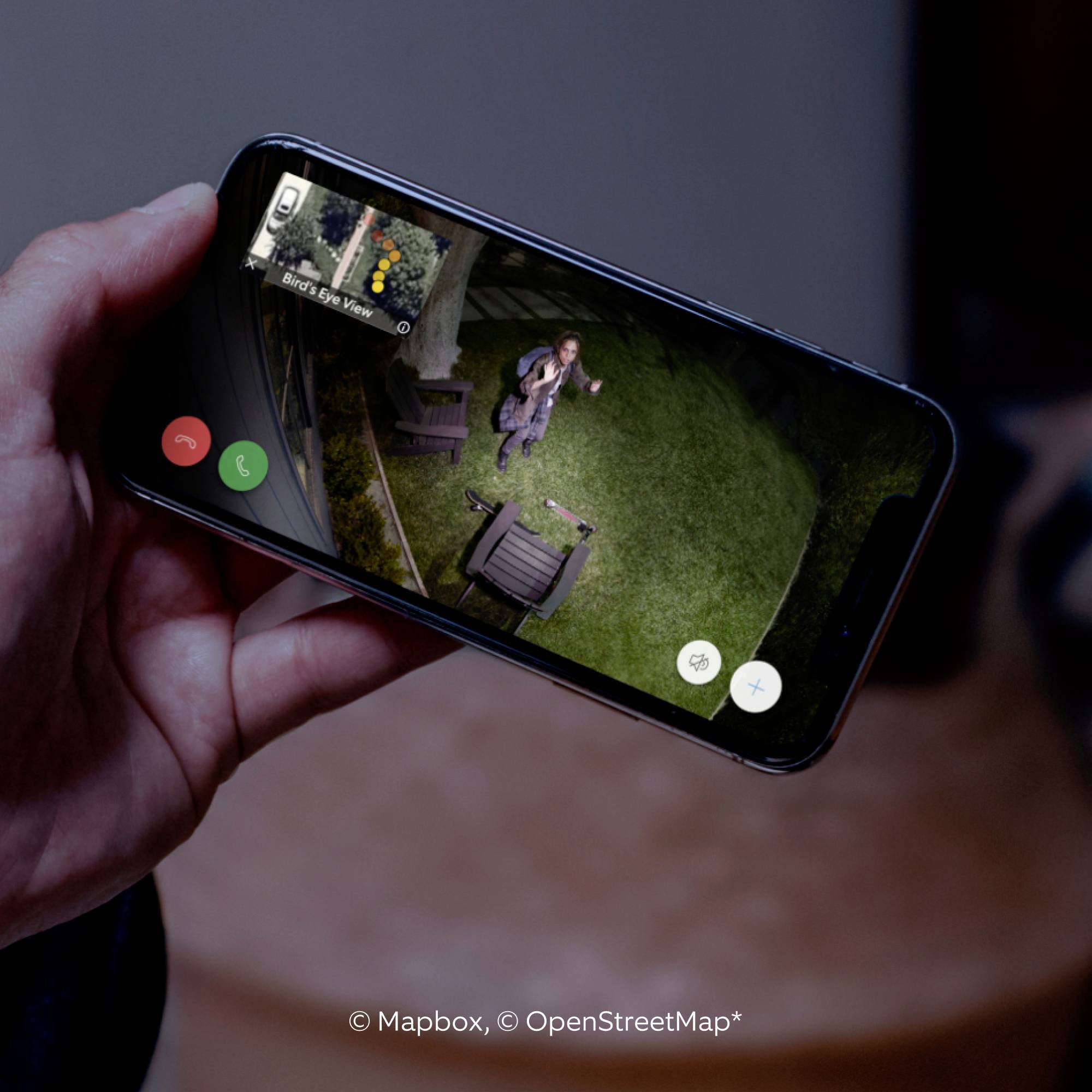 Jedna ruka drží smartphone, který zobrazuje letecký snímek muže v zahradě. Poskytovatelé map jsou 'Mapbox, OpenStreetMap'.