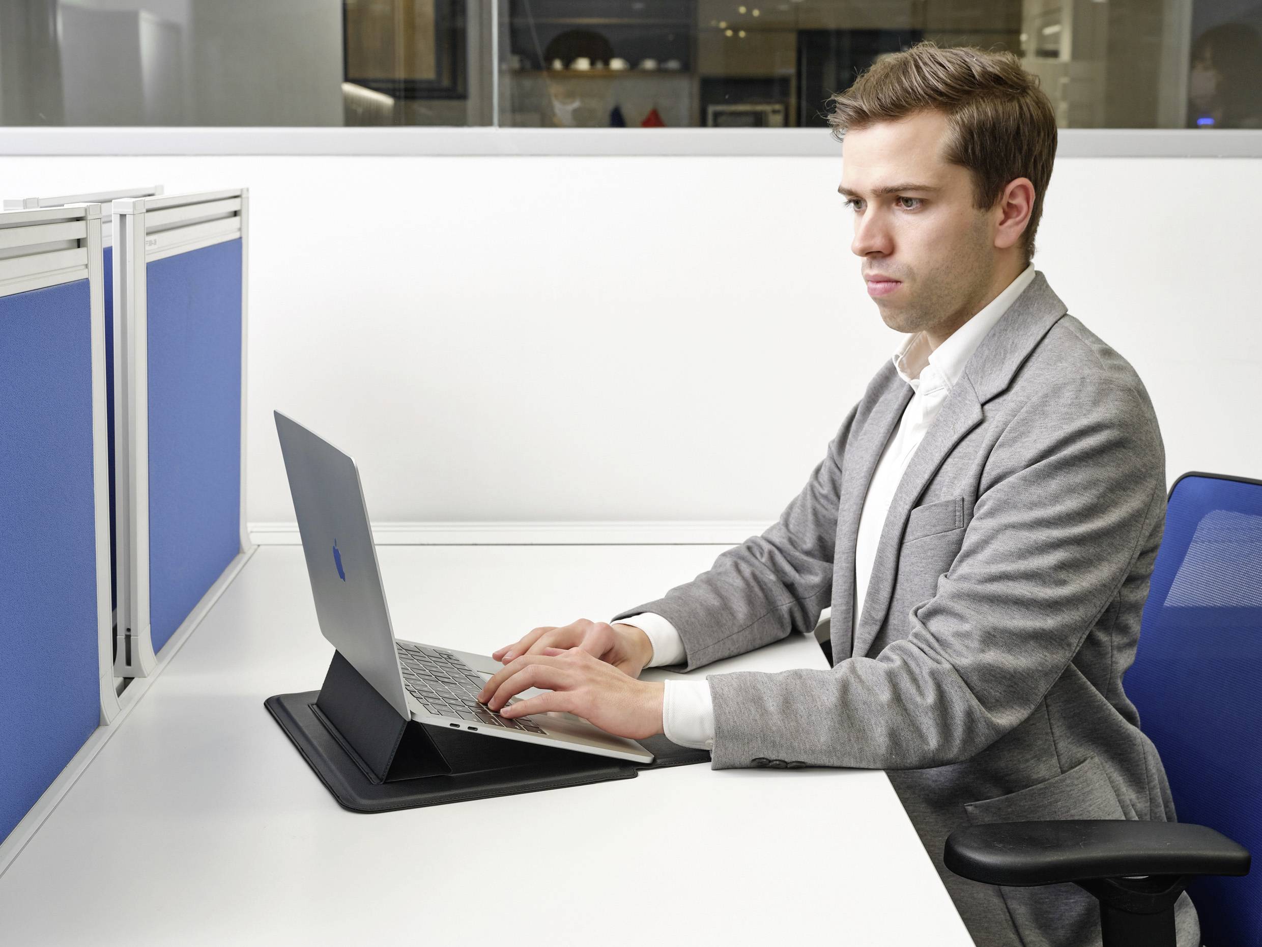 Muž v obleku sedí u psacího stolu a pracuje na laptoptpu. V pozadí jsou kancelářské stěny s modrými přepážkami.