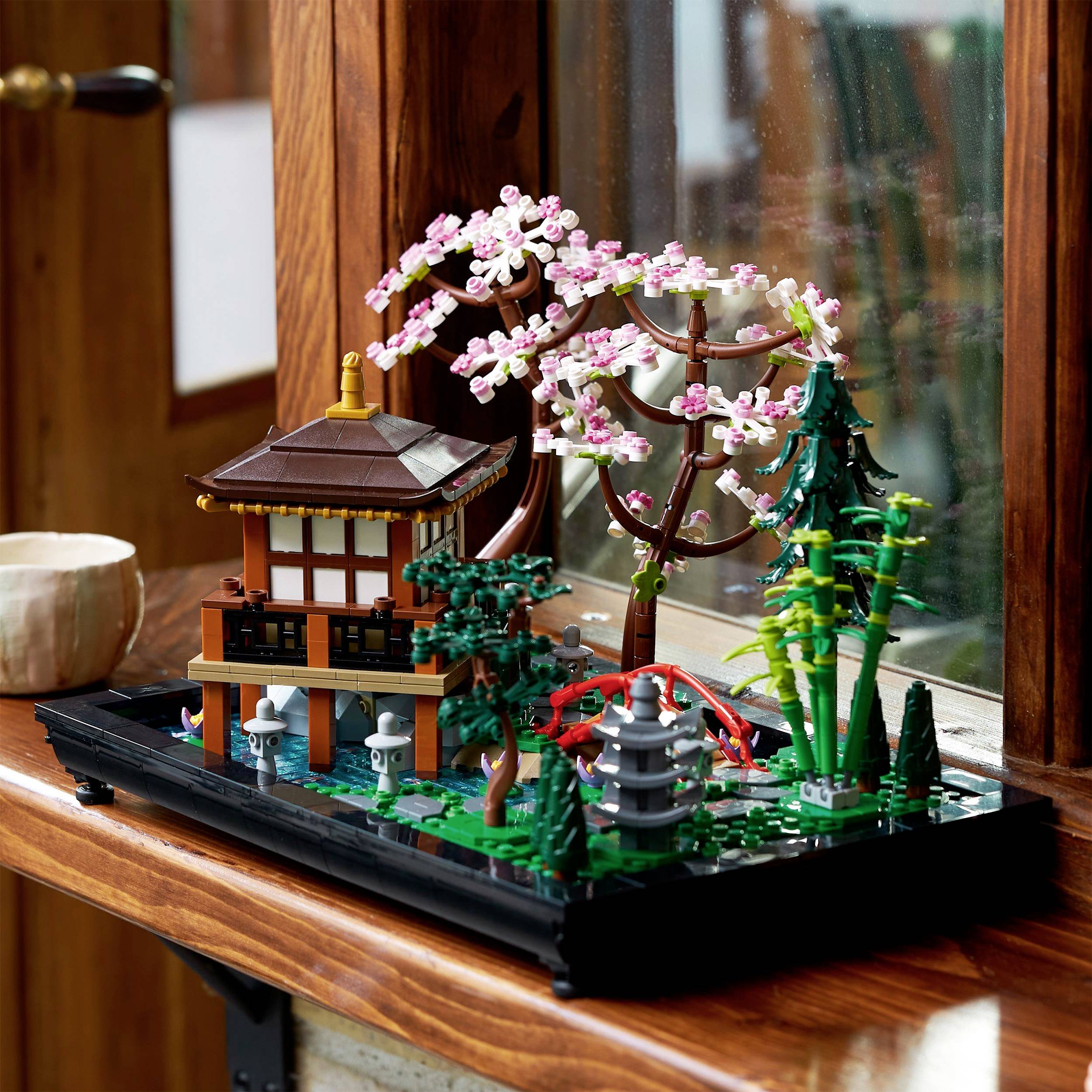 Detailní model japonské zahrady z LEGO kostek s pavilonem, kvetoucí třešní a mostem na dřevěném stole.