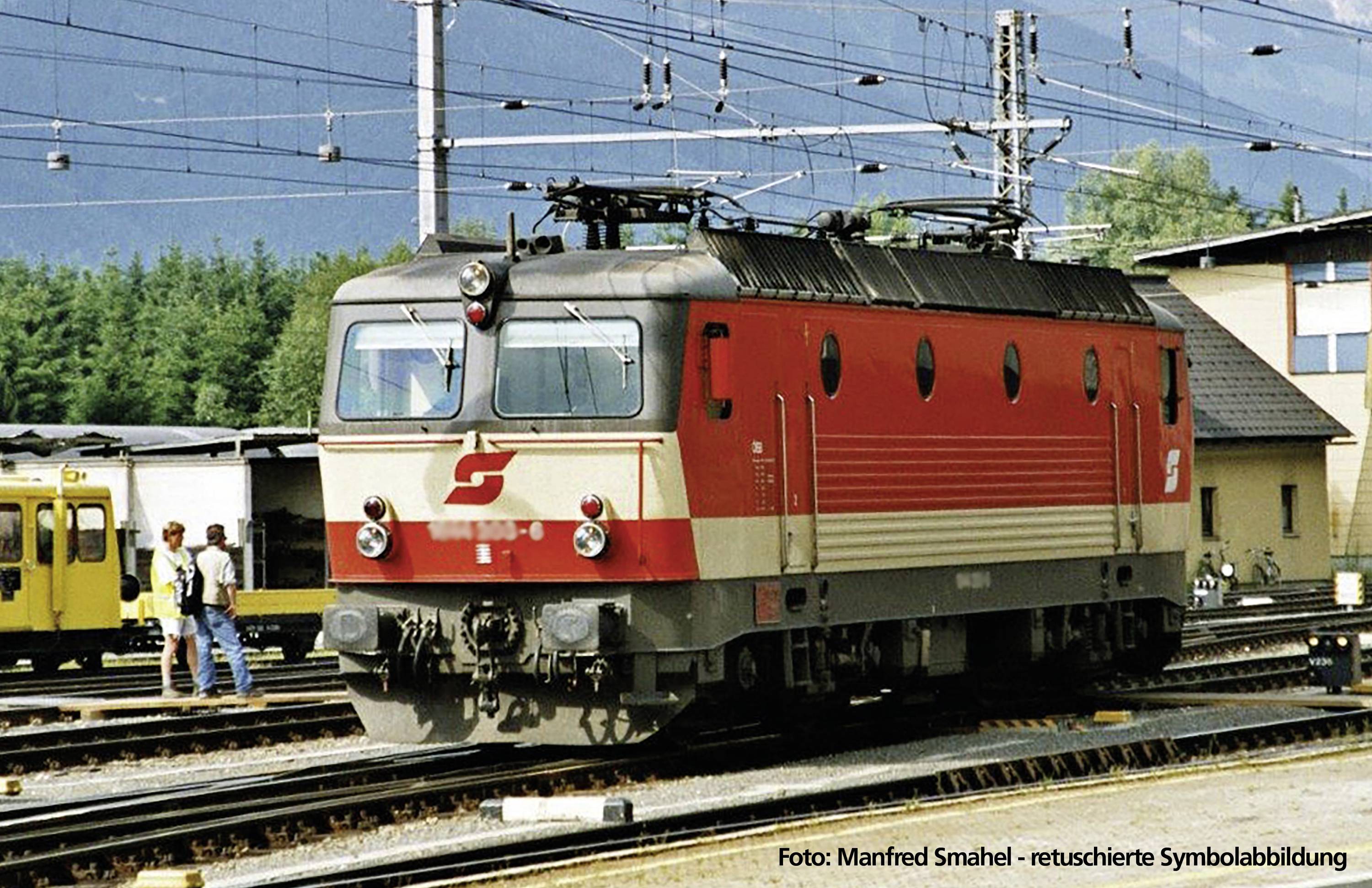 Červená a šedá elektrická lokomotiva stojí na koleji nádraží, obklopená dalšími kolejemi a budovami v pozadí.