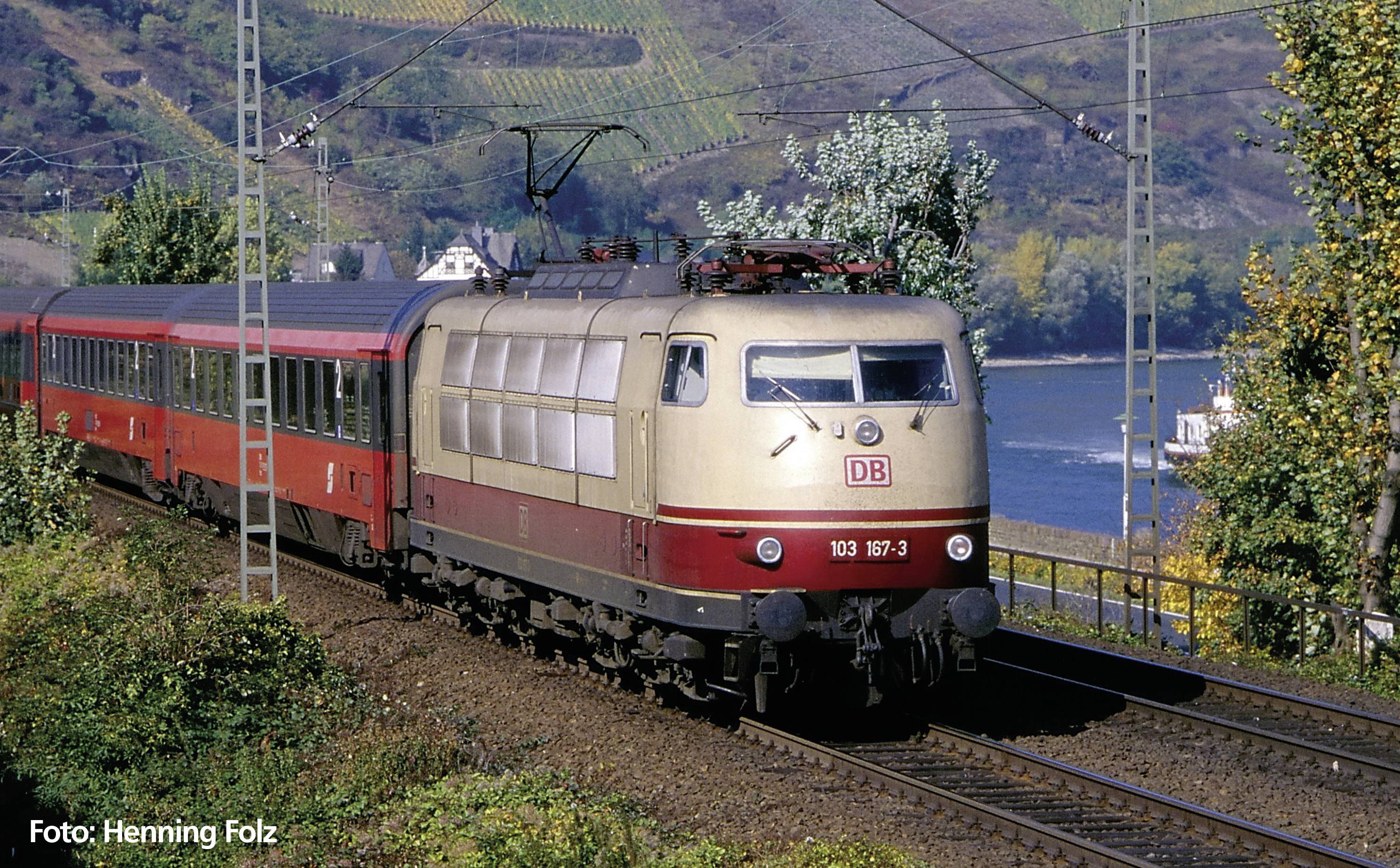 Červeno-béžový vlak Deutsche Bahn jede po trati v krajinné scenérii s kopci a stromy.