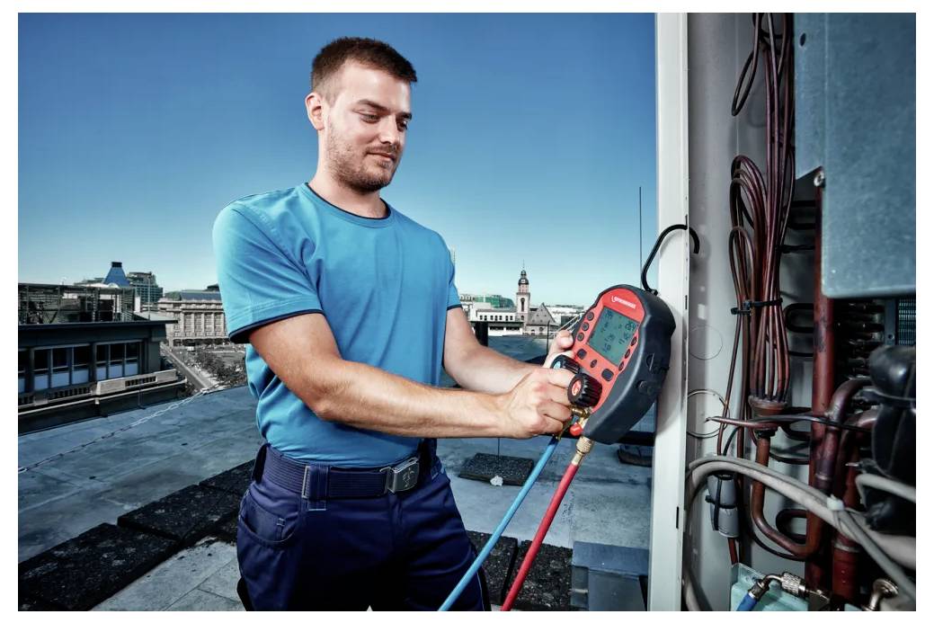 Technik v modrém tričku používá digitální multimetr k kontrole HVAC zařízení na střeše. V pozadí je vidět městská krajina.