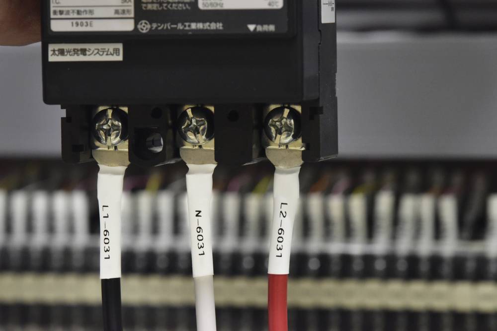 Černé zařízení se třemi připojenými barevnými kabely, označené štítky 'L1-6031', 'N-6031' a 'L2-6031'. V rozmazaném pozadí.