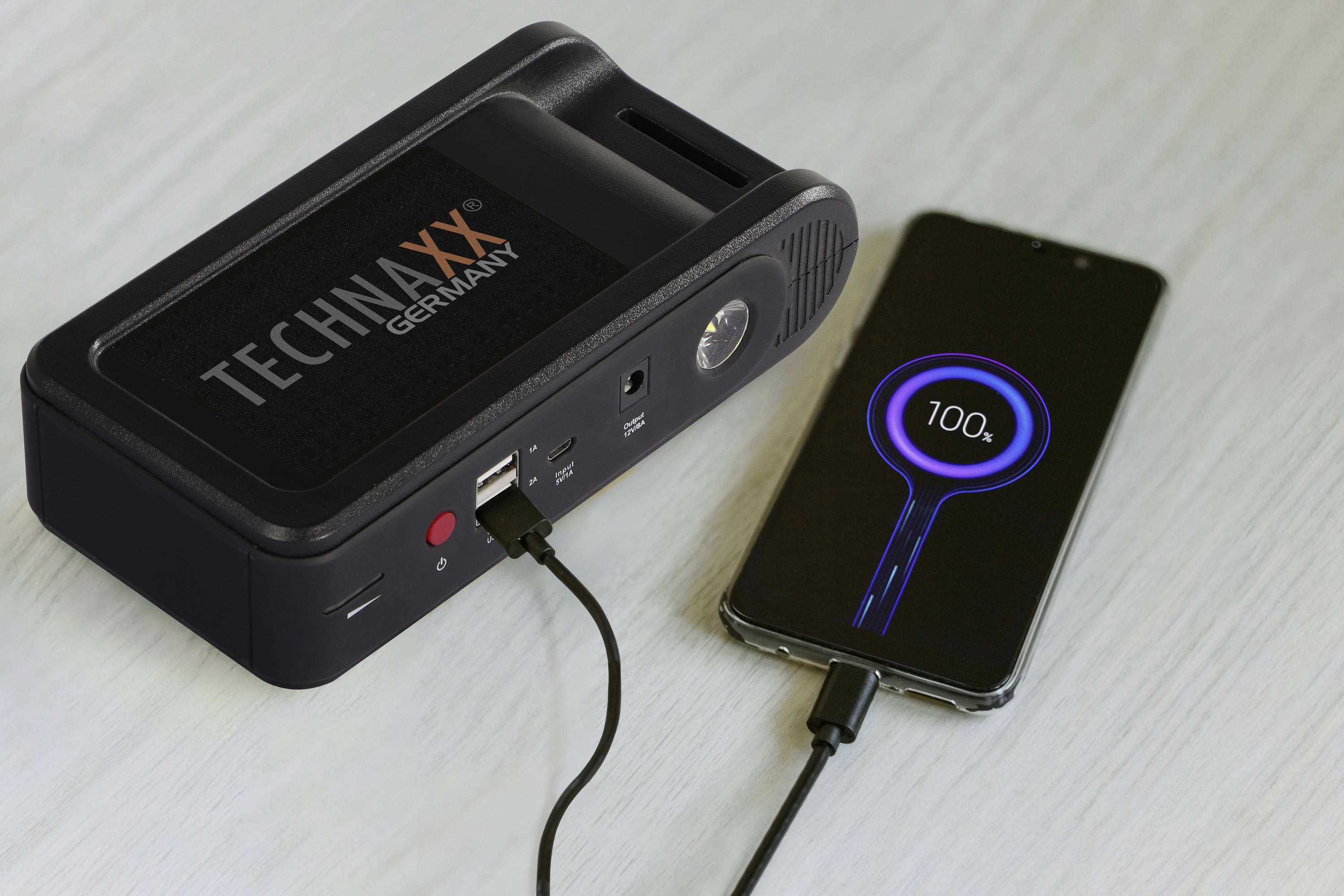 Smartphone je nabíjeno powerbанkou prostřednictvím kabelu. Display ukazuje 100% stav baterie.