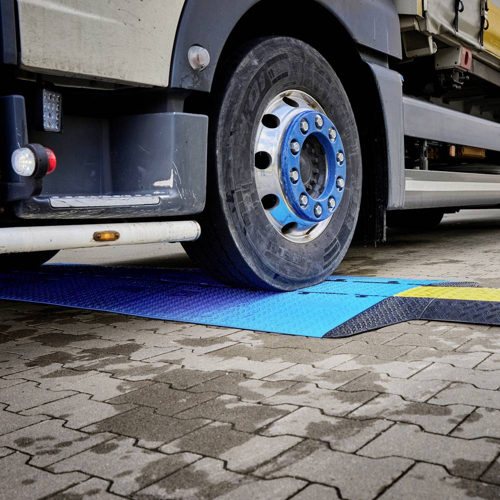 Nákladní automobil jede přes modrou váhu na dlážděné parkovací ploše. Váha měří hmotnost vozidla.