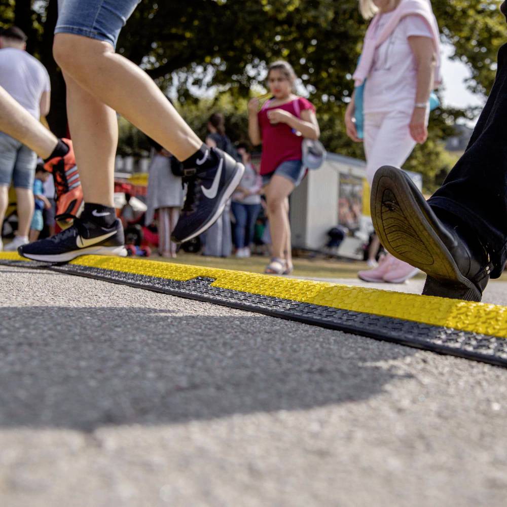 Lidé kráčí po chodníku v rušném prostředí, někteří mají sportovní obuv. V pozadí jsou viditelné stromy a budovy.