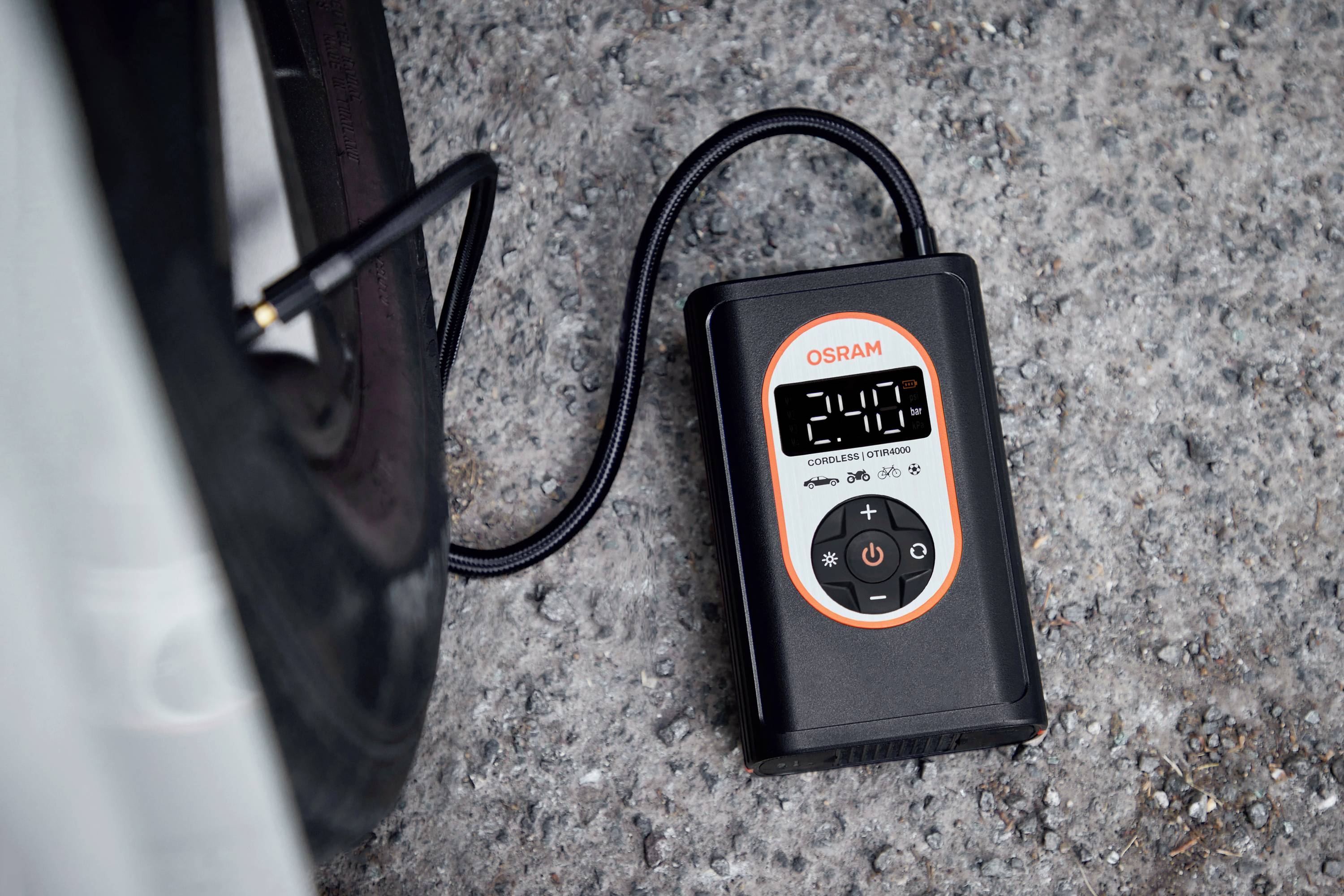 Digitální tlakoměr pneumatik ukazuje '2.4 Bar' a je připojen k ventilku automobilové pneumatiky. Zařízení leží na štěrku.