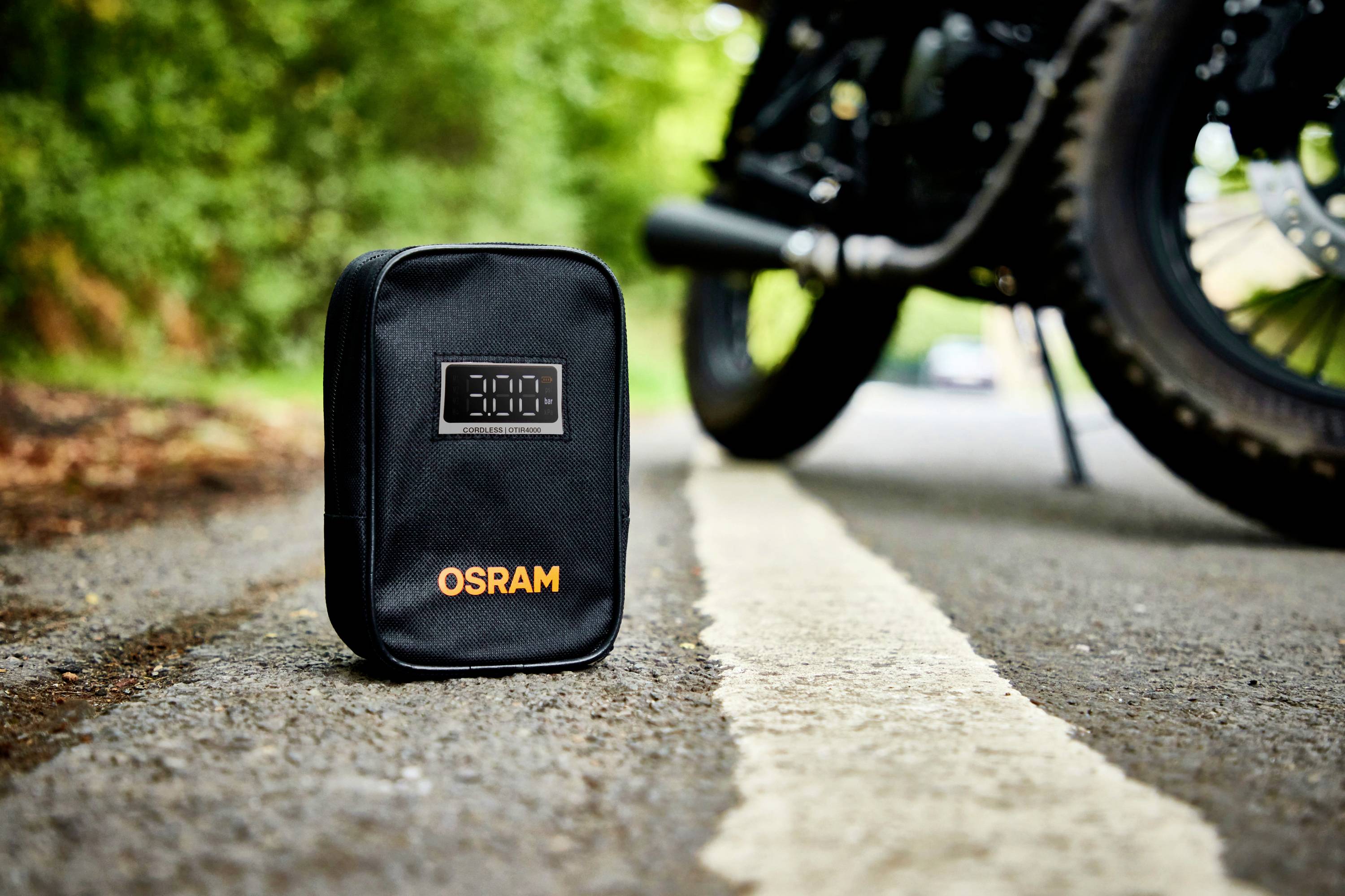 Černá taška s potiskem 'OSRAM' stojí na silnici vedle motocyklové pneumatiky. V pozadí je viditelná zelená vegetace.