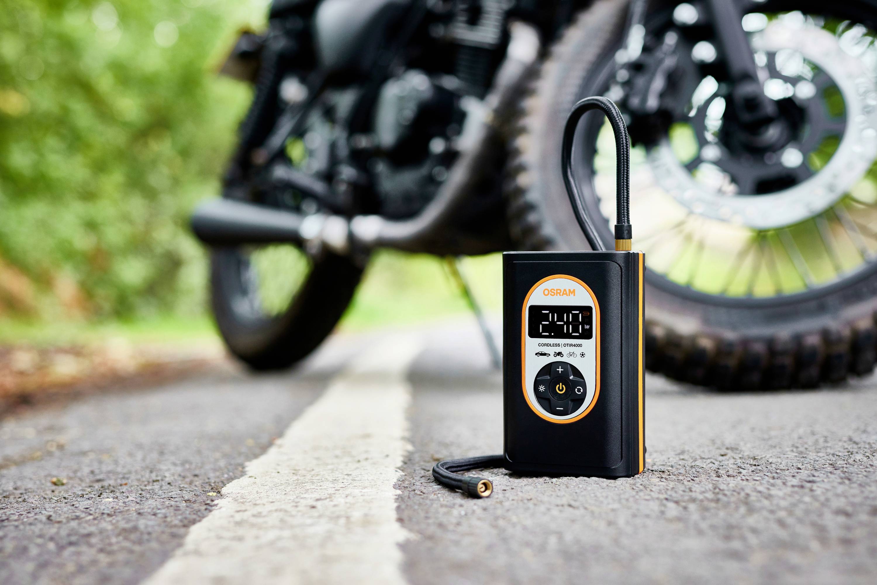 Přenosný kompresor na pneumatiky ukazuje '2,40' a stojí na silnici před motocyklem.