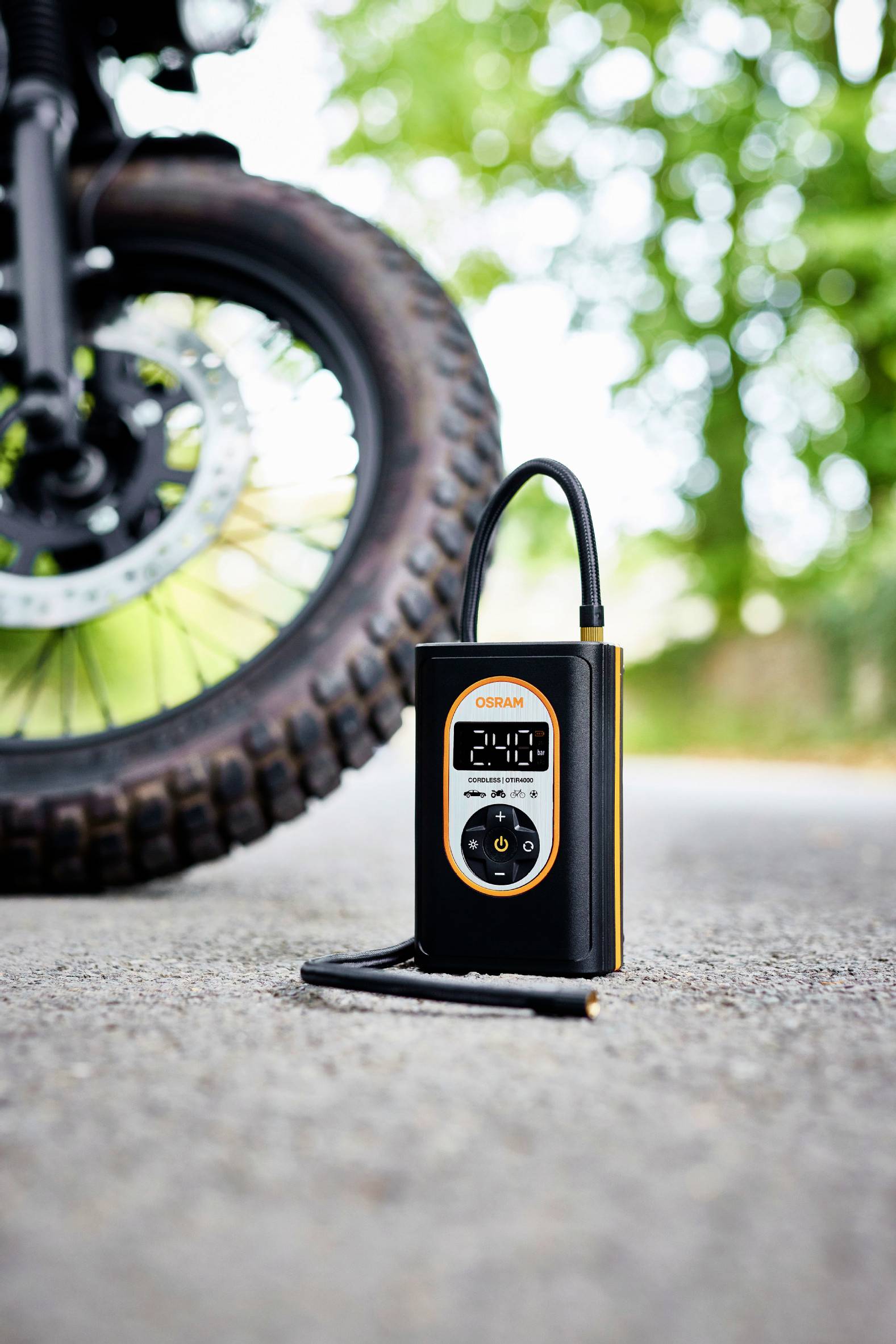 Přenosný měřič tlaku v pneumatikách na asfaltu vedle motocyklové pneumatiky, venku před stromy.